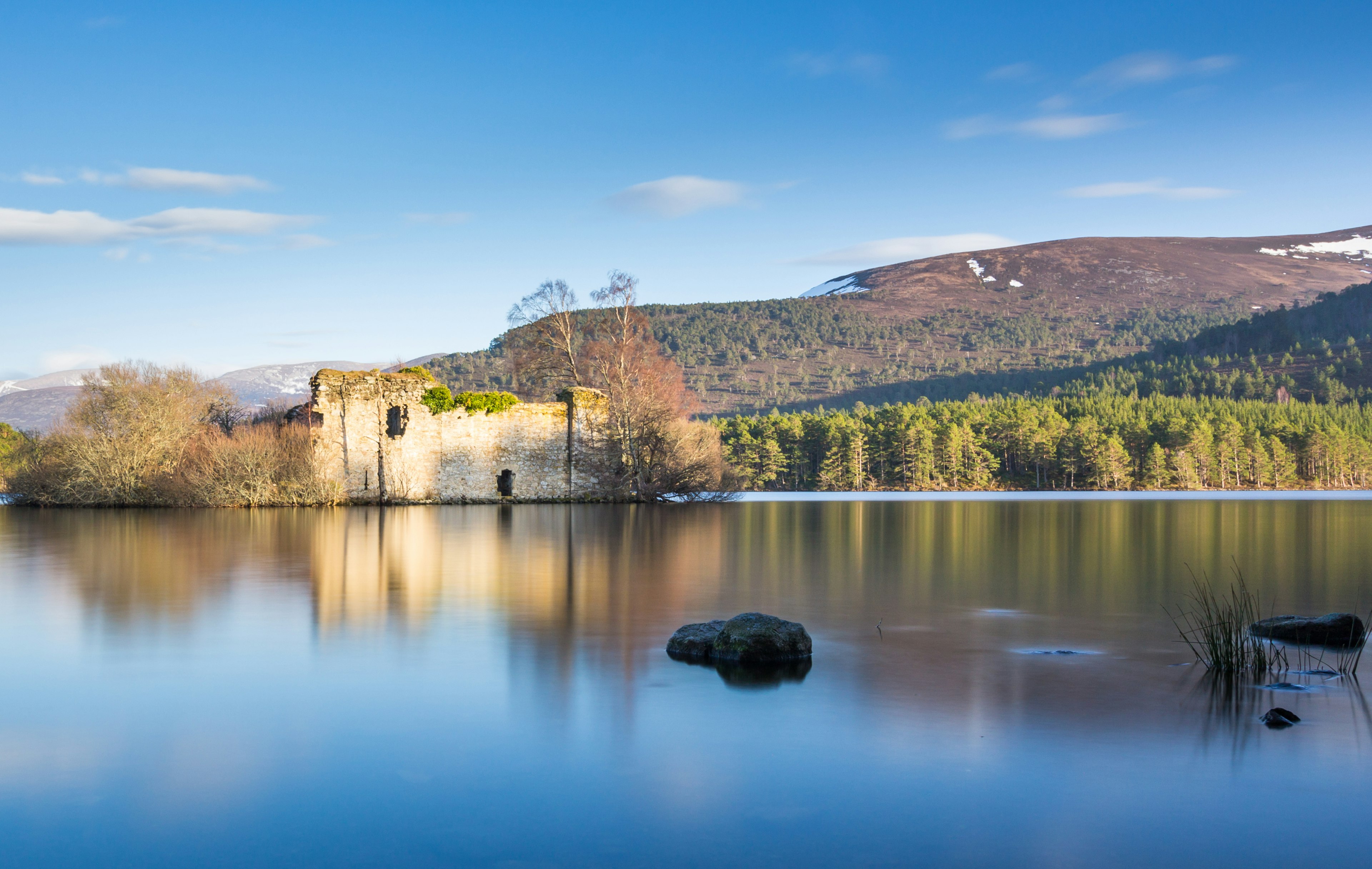 A view from the banks of Loch An Eilein, looking out towards the ruined castle on an island in the middle of the Loch. Beyond, the opposite forest-lined bank is visible, leading away to rolling mountains.