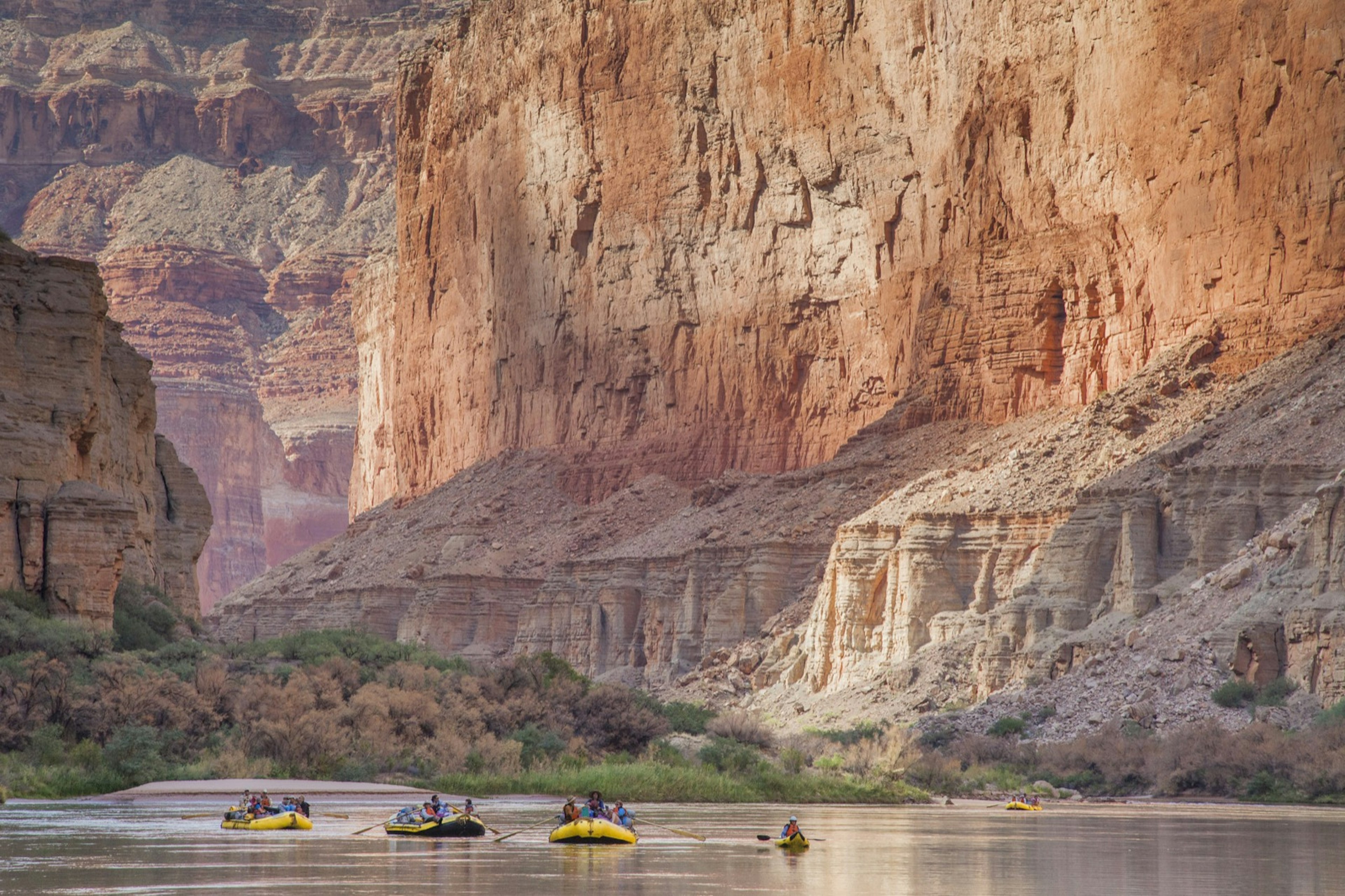 Whitewater rafts and a kayak drift on a calm part of the Colorado River in the Grand Canyon, as sheer rock walls fill the frame behind.