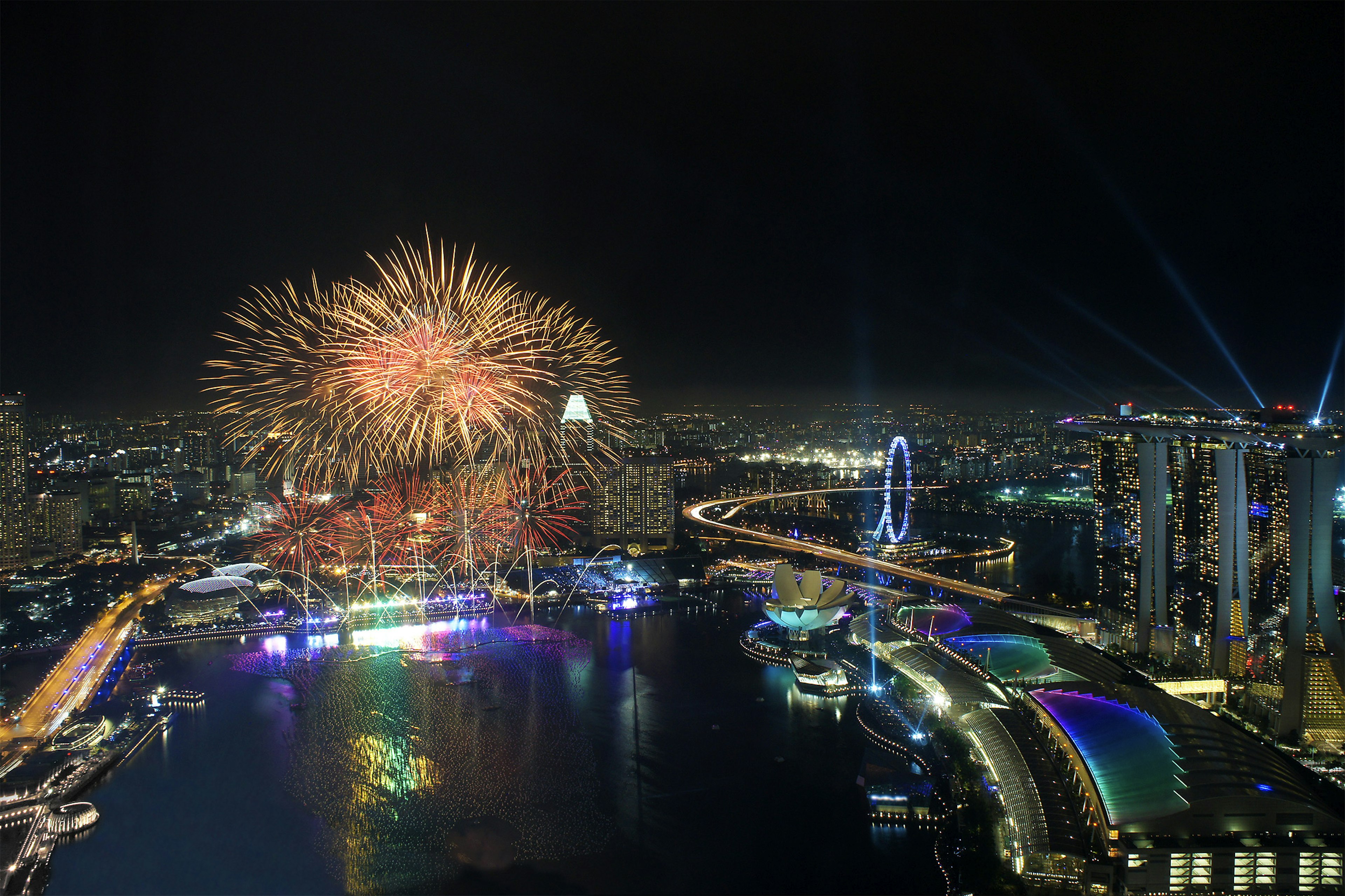 An aerial view of the Singapore NYE firework display. With a series of bright fireworks lighting up the sky above the city's skyscrapers.