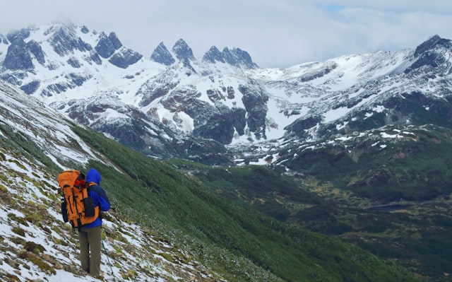 A hiker on a steep slope with snow on green vegetation. Sharply peaked, snow-covered mountains are in the distance. Dientes de Navarino Circuit, Isla Navarino, South Chile