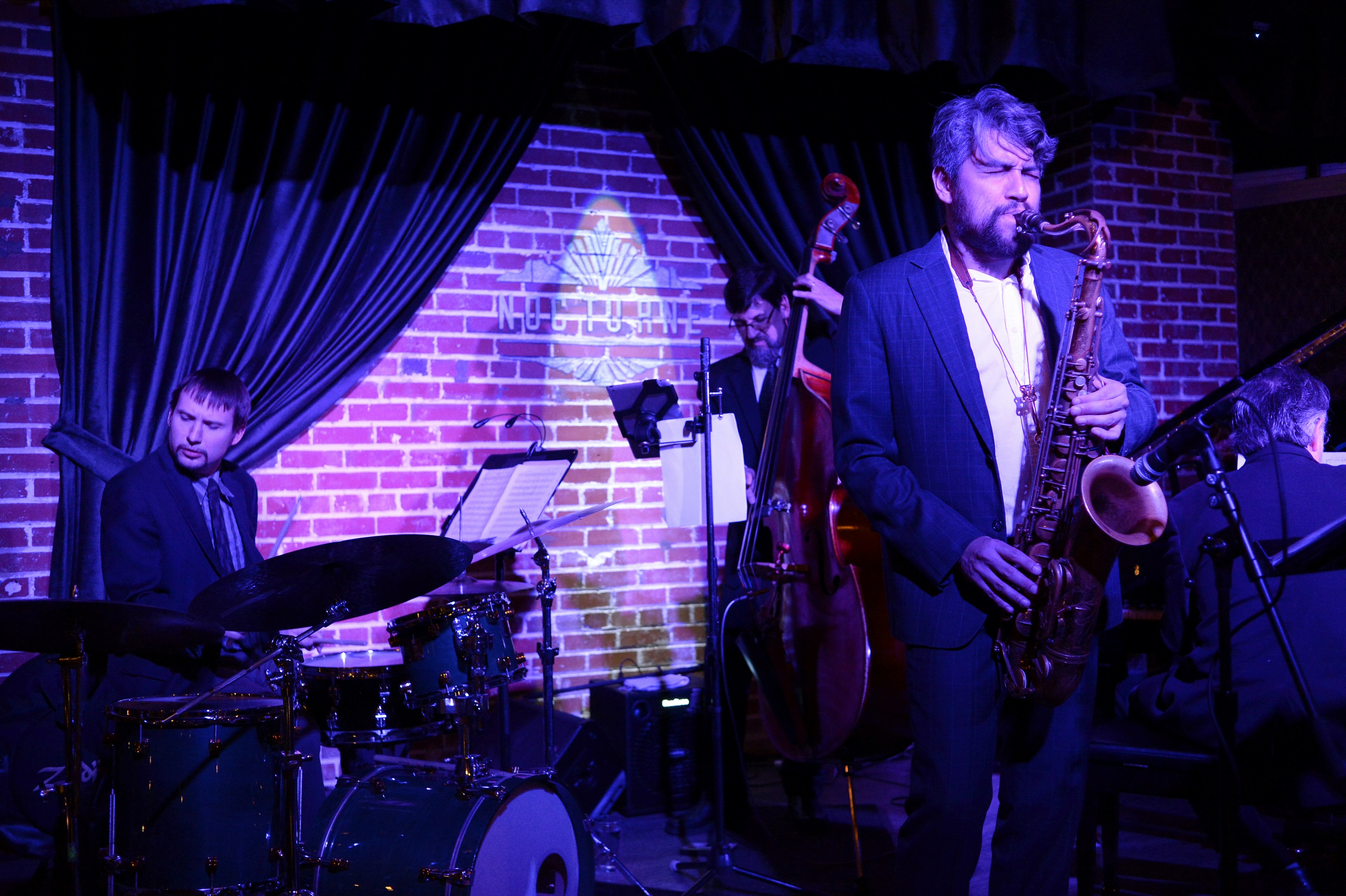 A jazz trio in dark casual suits play an upright bass, a saxophone, and drums under purple lights on stage at Nocturne jazz club