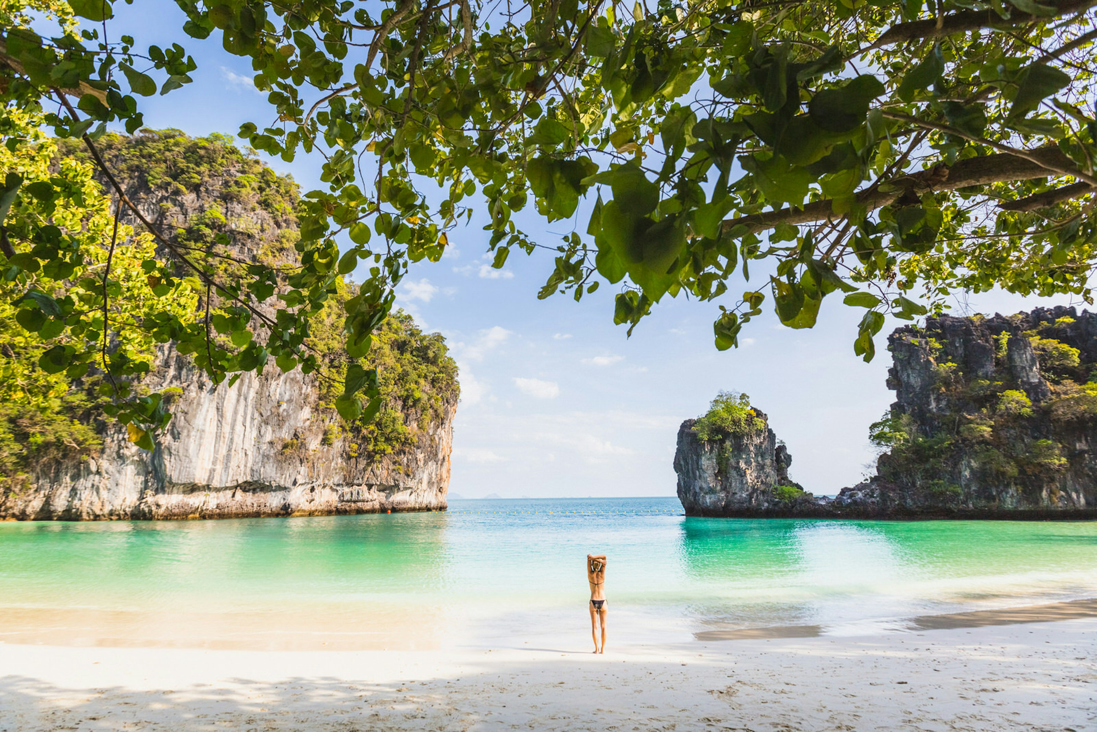 A woman wearing a bathing suit stands on the shore of a beach looking at large green-covered rocks coming out of the ocean water