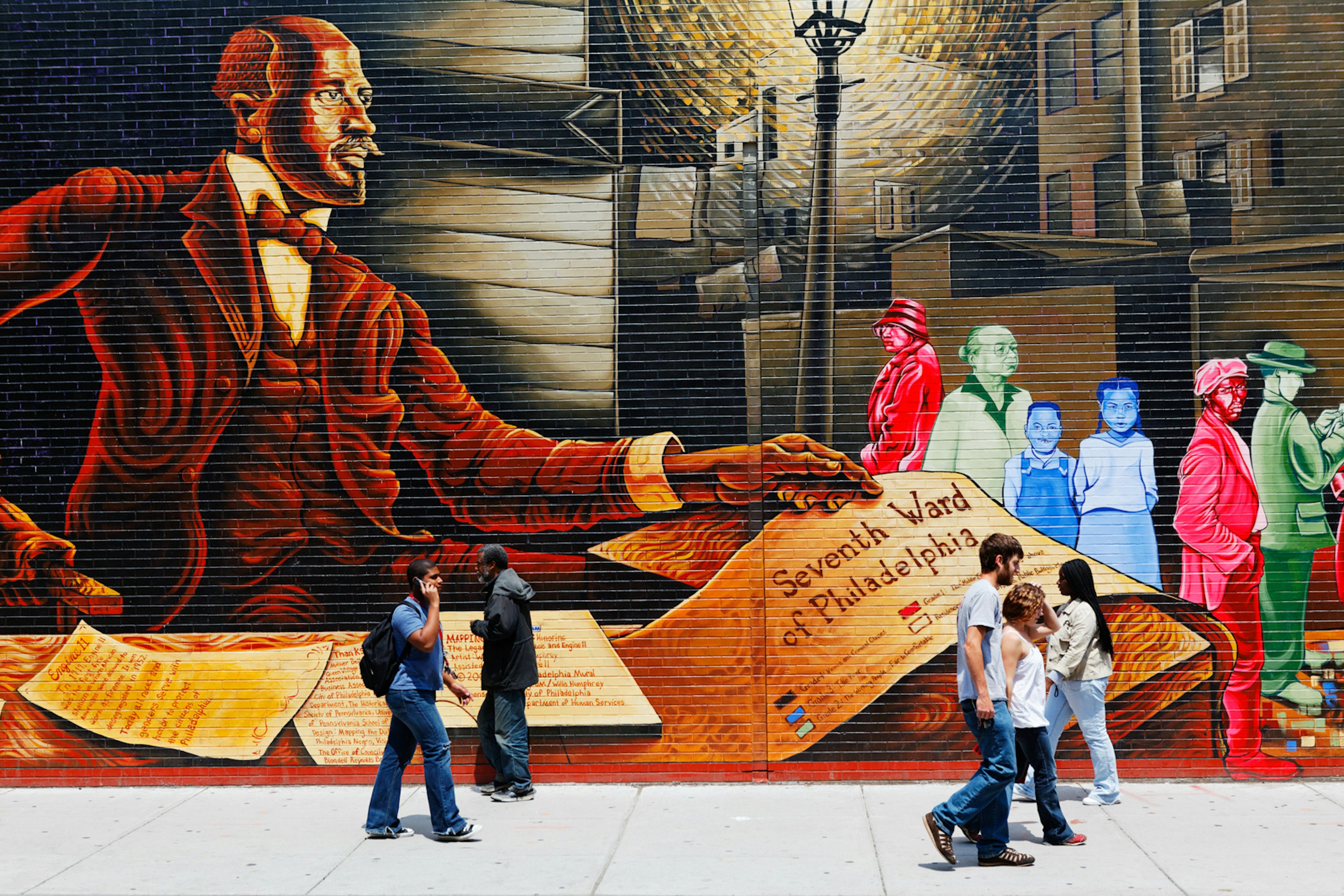 A colourful wall mural depicting W.E.B Du Bois, with people on the sidewalk nearby. The painting shows an oversized man at a desk littered in papers, with smaller people painted in red, green and blue to the right of him standing on a street under a light.