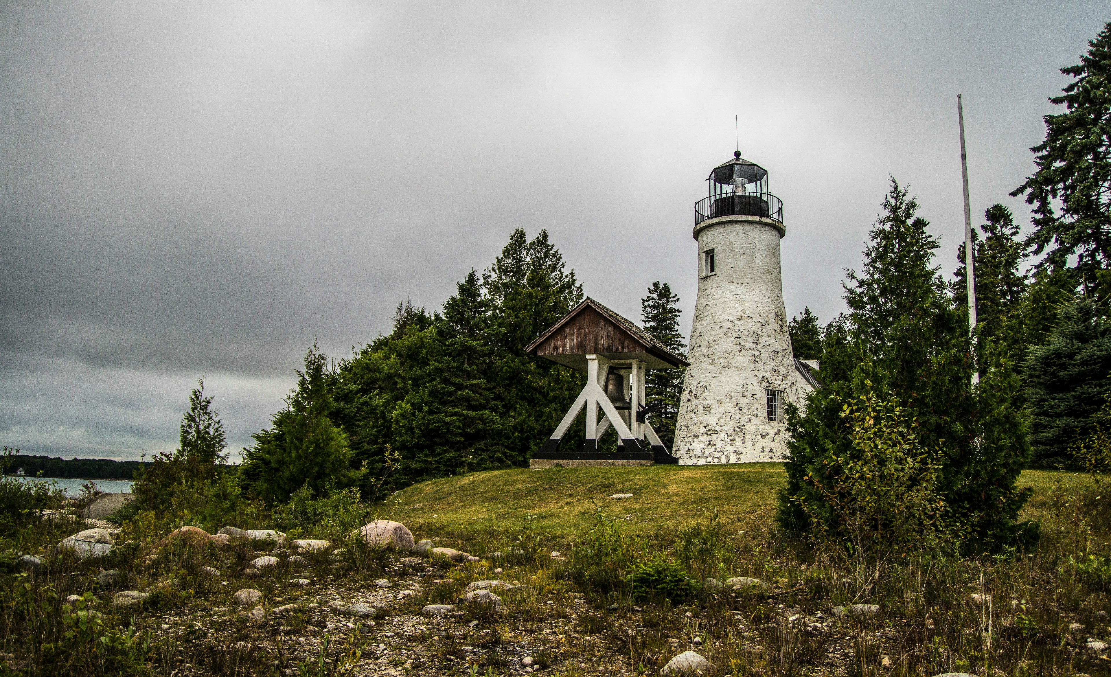 The exterior of the Old Presque Isle Lighthouse on the coast of Lake Huron. A white, round brick tower with a metal railing on top around the glass panes surrounding the light itself. Small rectangular windows are cut in on the lower right and upper left of the structure. To the left of the lighthouse is a short white wooden belfry with an enormous metal bell. All around is the rocky coast and bright green trees.