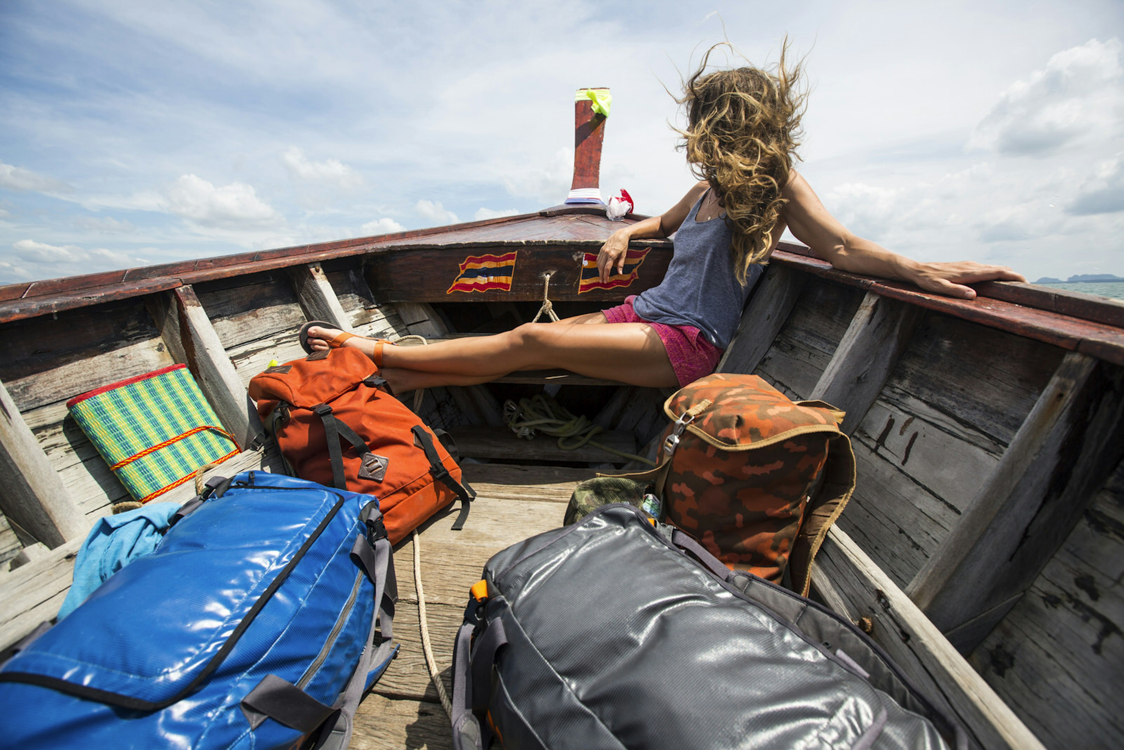 A woman sits at the front of a boat with her legs up as the sea breeze blows through her hair. Beside her are two small book bags and two larger duffle bags.