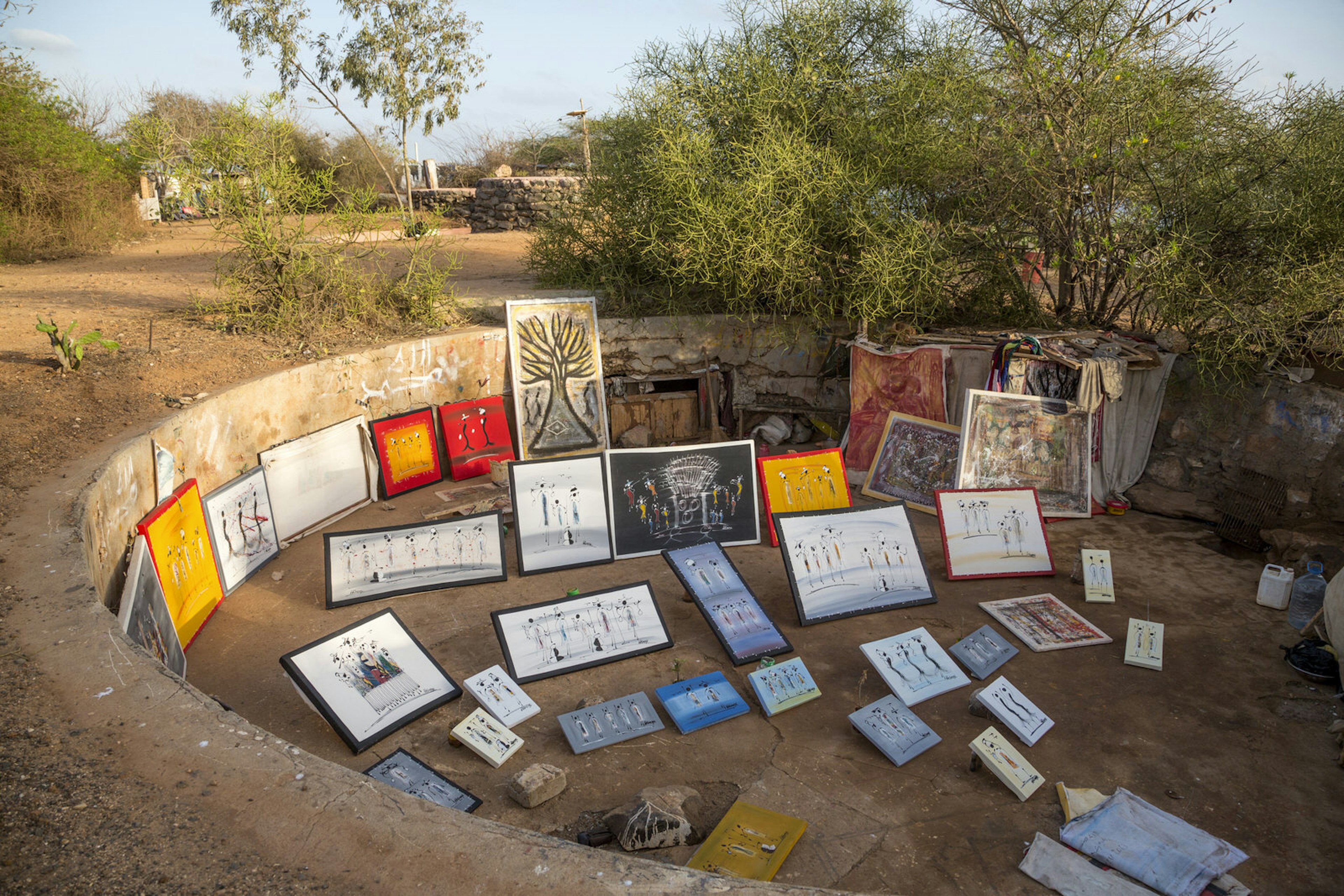 Within the open-air semi-circular gun bunker are dozens of colourful paintings - either on the dirt ground or balanced against the cement wall. Vegetation grows in the surrounding area © Rhapsode / Getty Images