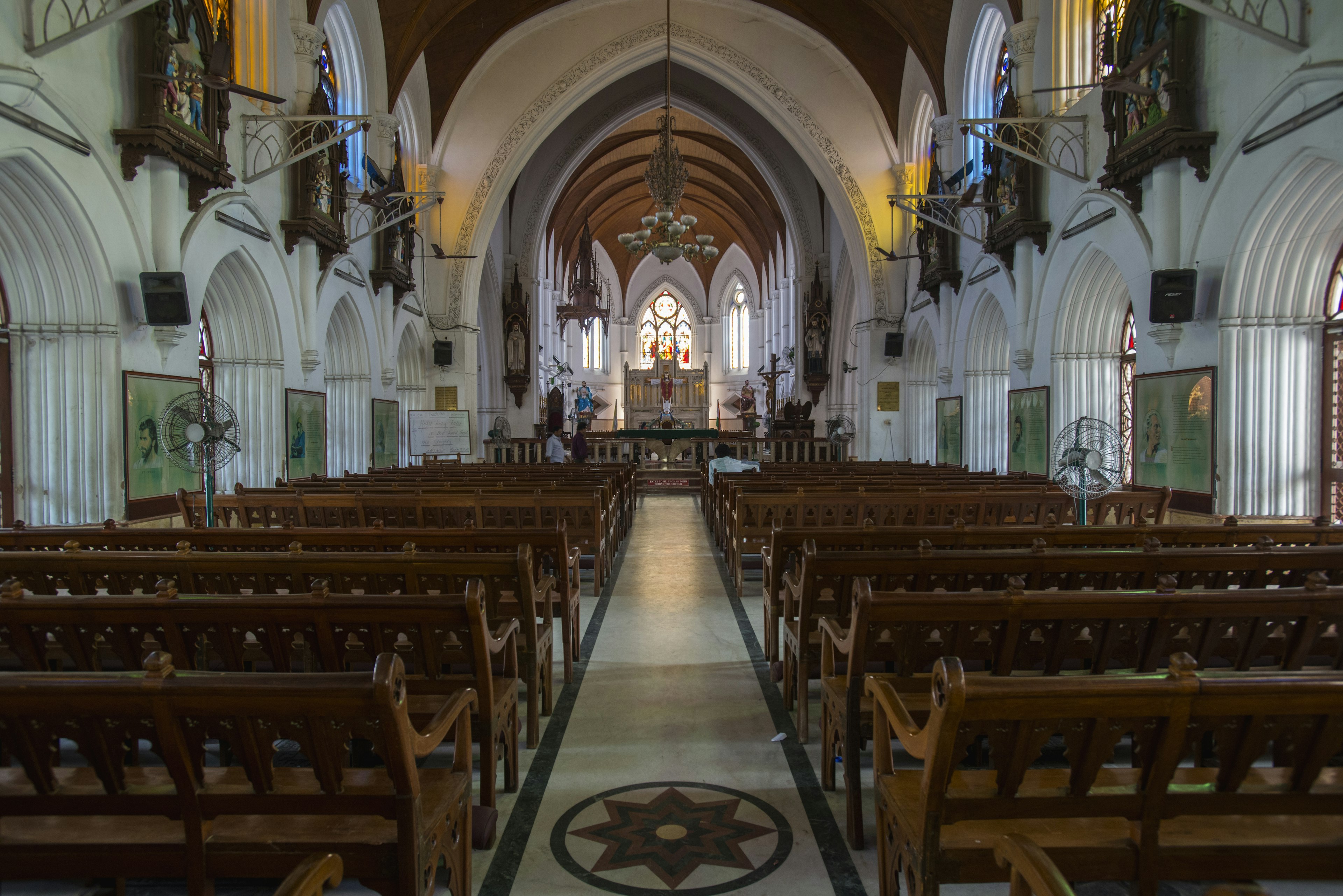 Interior of a cathedral, looking directly down the central aisle. There are dark wooden pews on either side and grand white arches overhead.