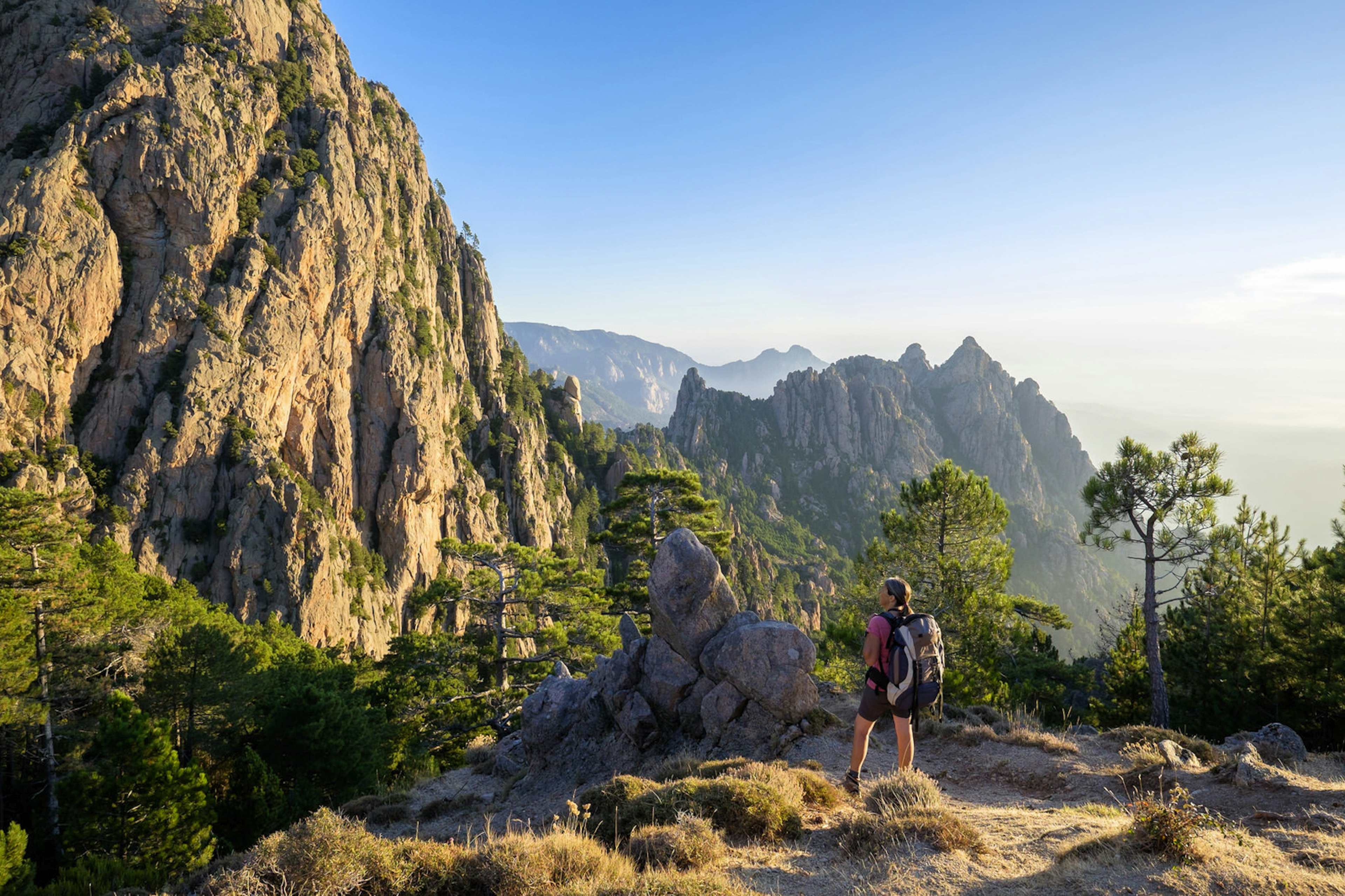 A woman with a backpack stands on a hilltop along the GR20 trail with rocky mountain peaks visible beyond her