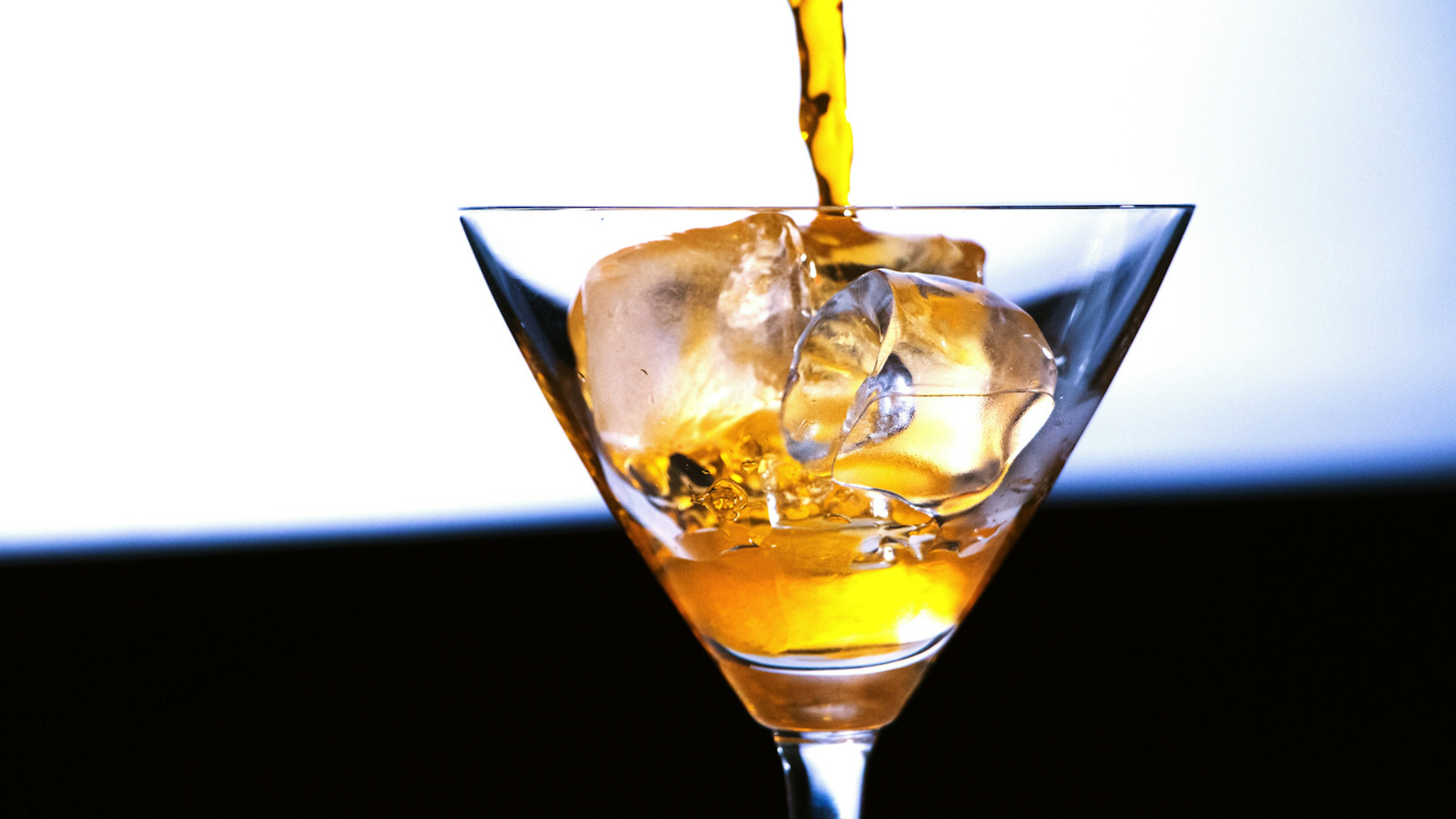 A golden-coloured cocktail being poured into a Martini glass against a white background