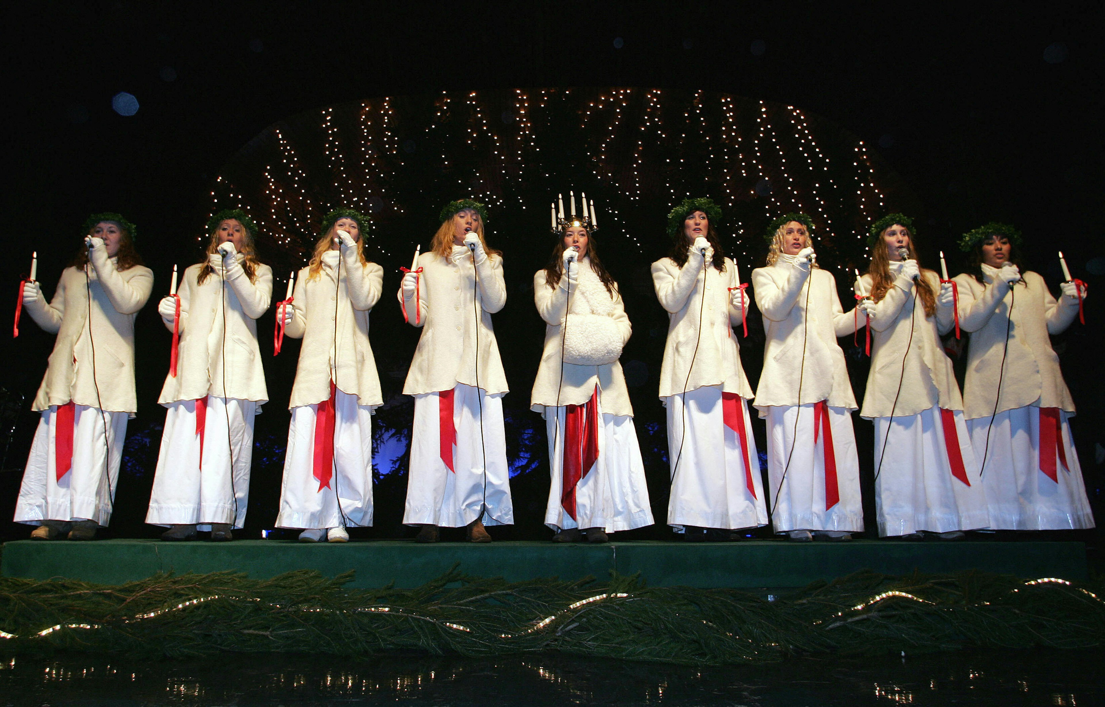A line of women in white dresses and wreaths on their heads singing on a stage on St Lucia's Day.
