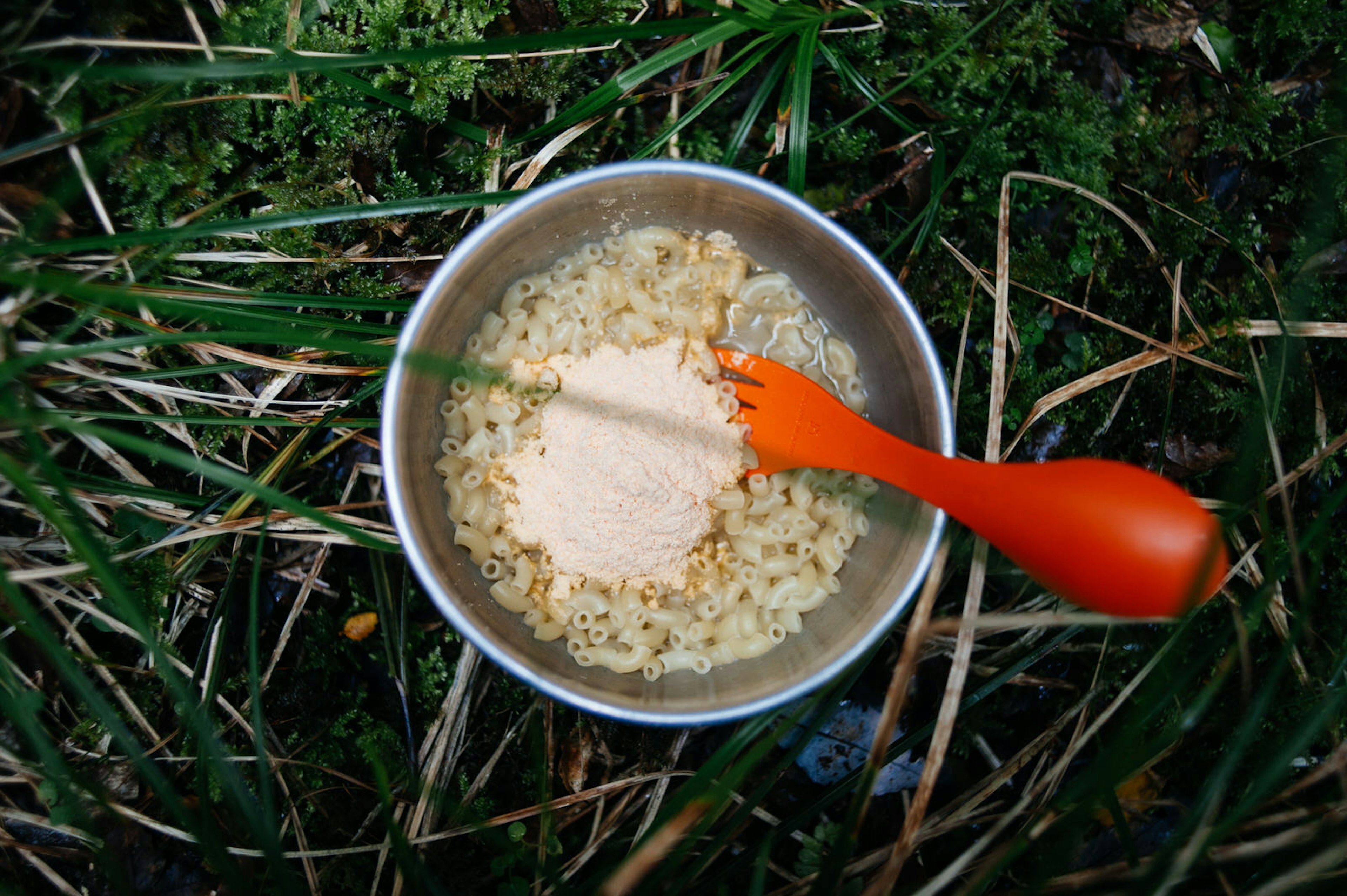 An orange spork in a pan of macaroni and powdered cheese sitting on some grass