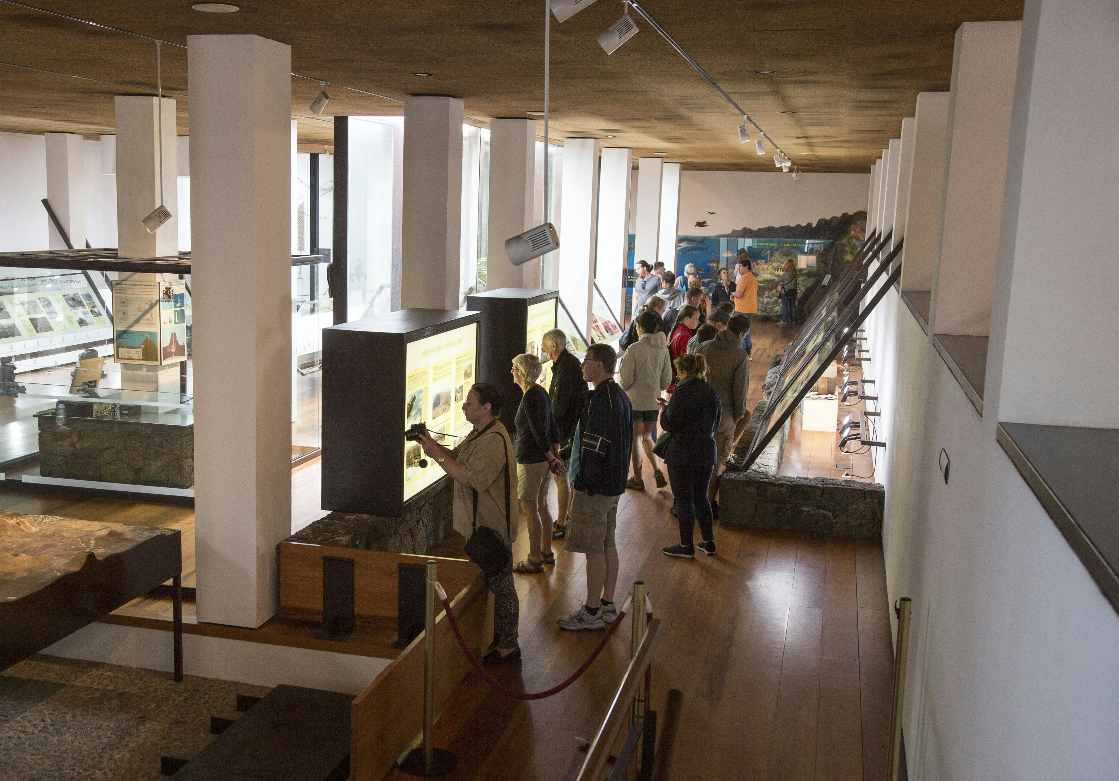 A group of tourists read visual displays and even photograph signage at Timanfaya Volcano Interpretation and Visitors Centre at the Canary Islands in Spain. The room in which the tourists stand is made up of white rectangular columns and smooth white plaster walls with a warm-toned brown ceiling and wooden floor with wide planks. The columns divide the room into two sections full of different exhibits and back-lit informational stations.