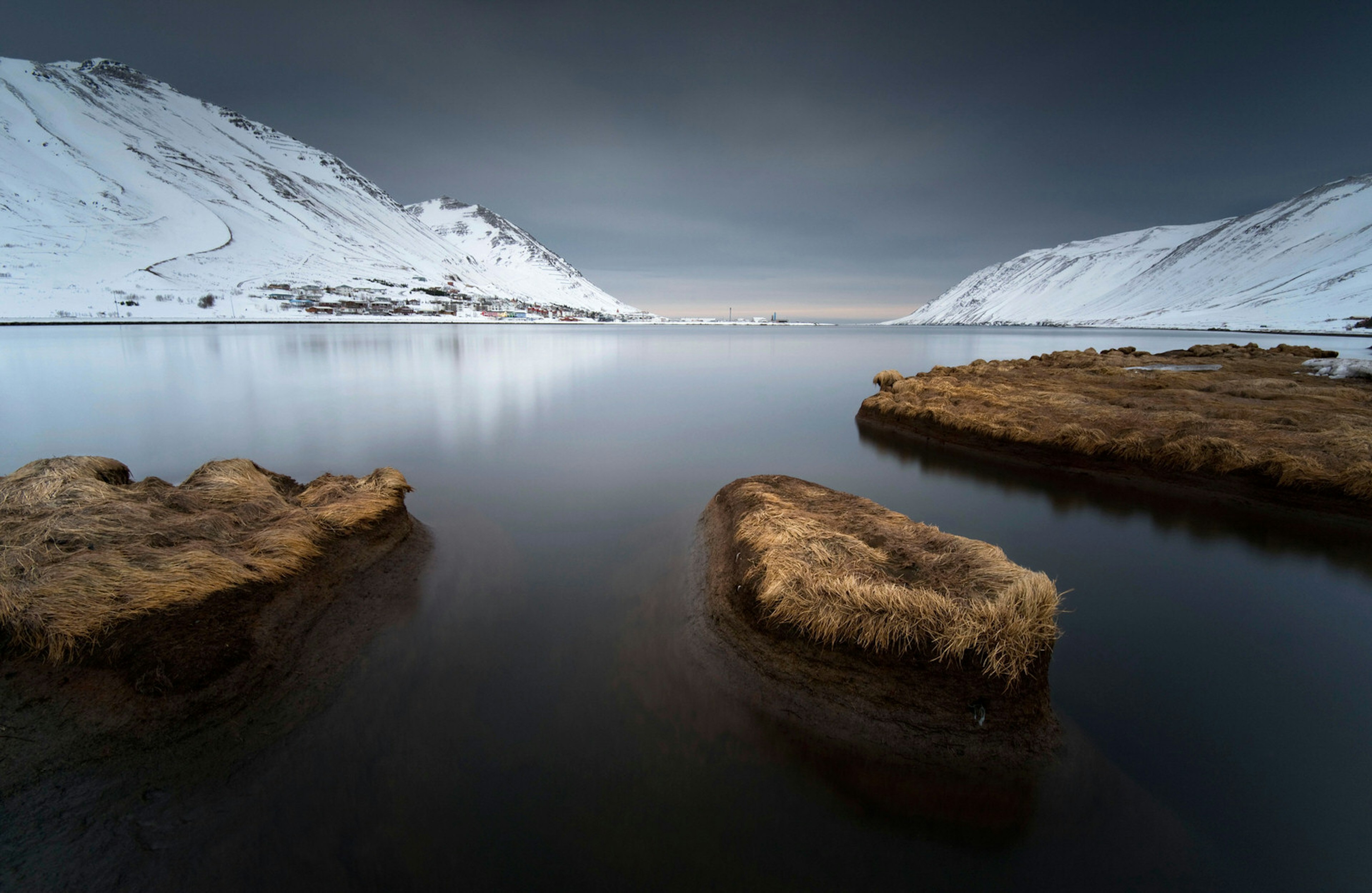 Snow-covered cliffs slope down into a perfectly still fjord