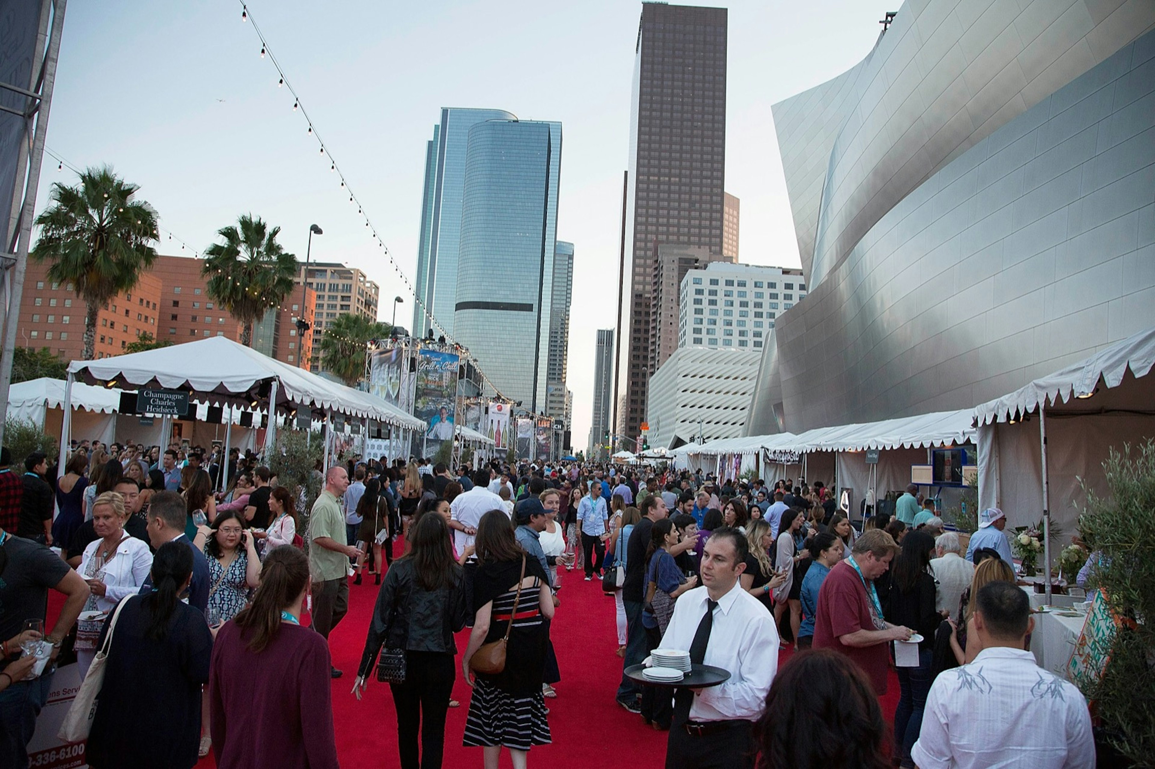 Well-dressed people mingle on a red carpet with skyscrapers in the background while a man in a tie holds a tray at one of California's best summer food and drink festivals