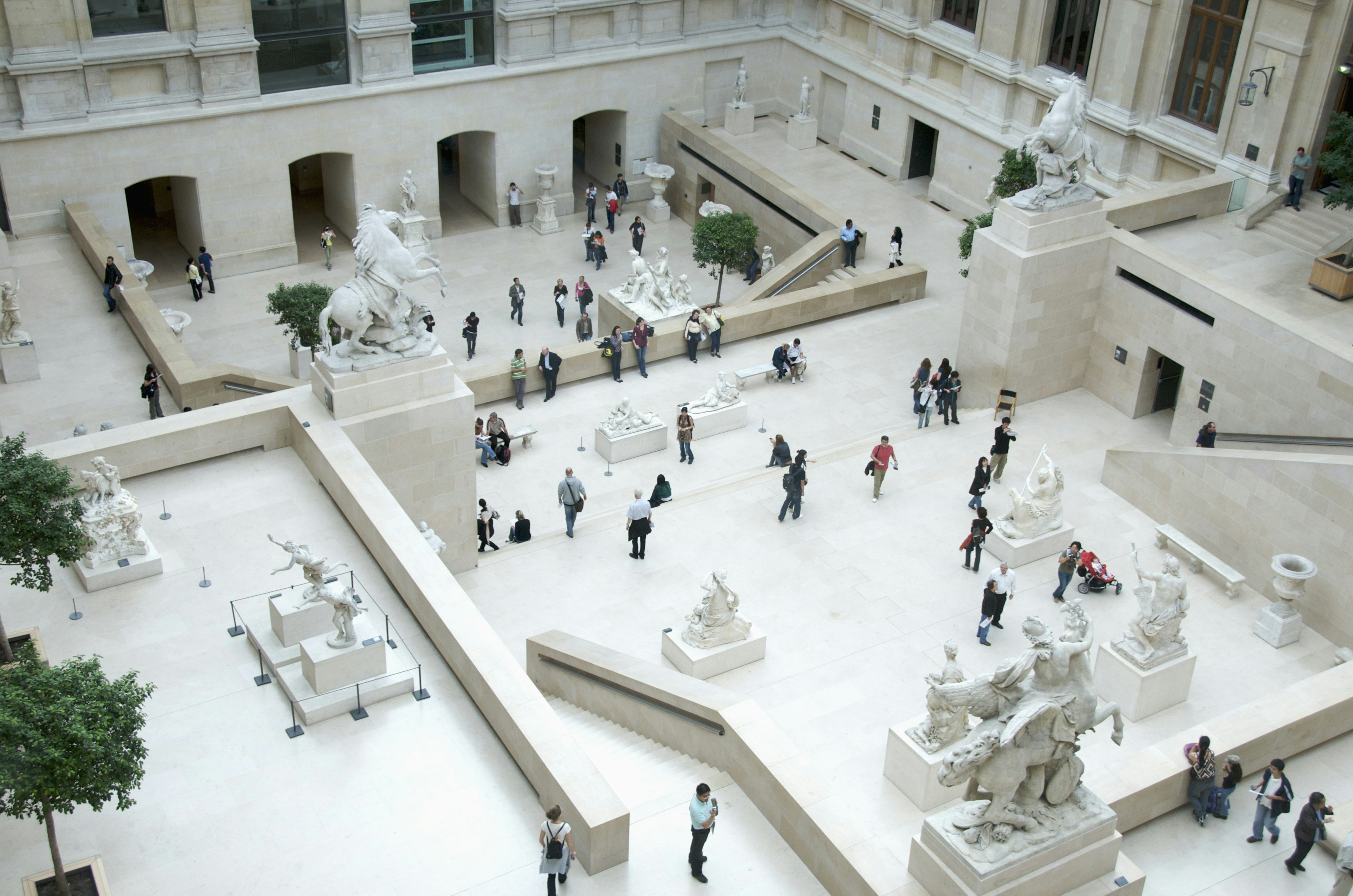 Aerial view of the Cour Marly, a courtyard in the Louvre filled with sculptures from the 17th and 18th centuries. The white courtyard is filled with visitors looking at the exhibits.