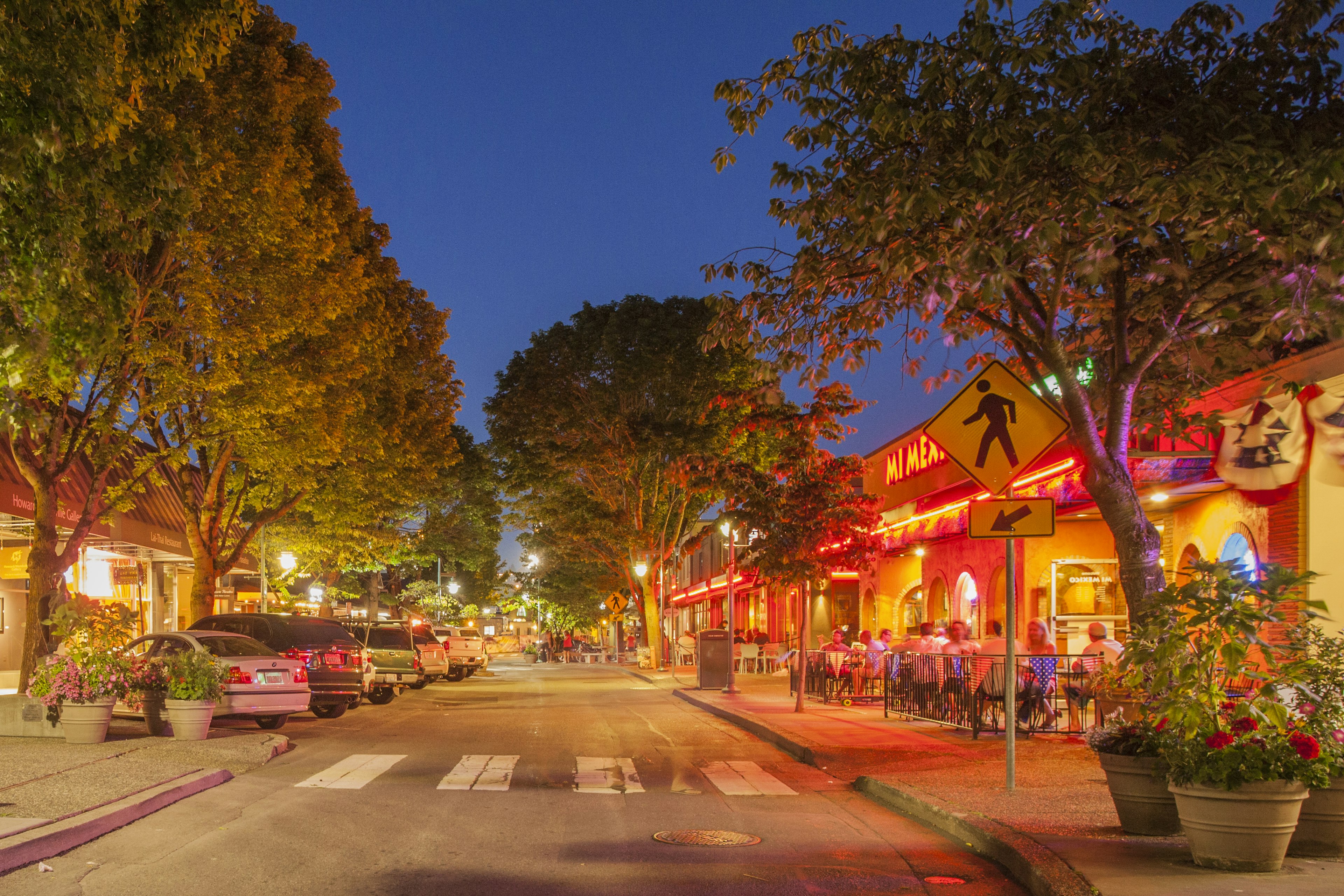 The orange and red light cast by neon storefront and restaurant signs on a street in downtown Kirkland, Washington illuminates big, full green trees and contrasts with a deep blue summer night sky. On the left side of the street is a row of parked cars. On the right, diners sit at cafe tables on the sidewalk.