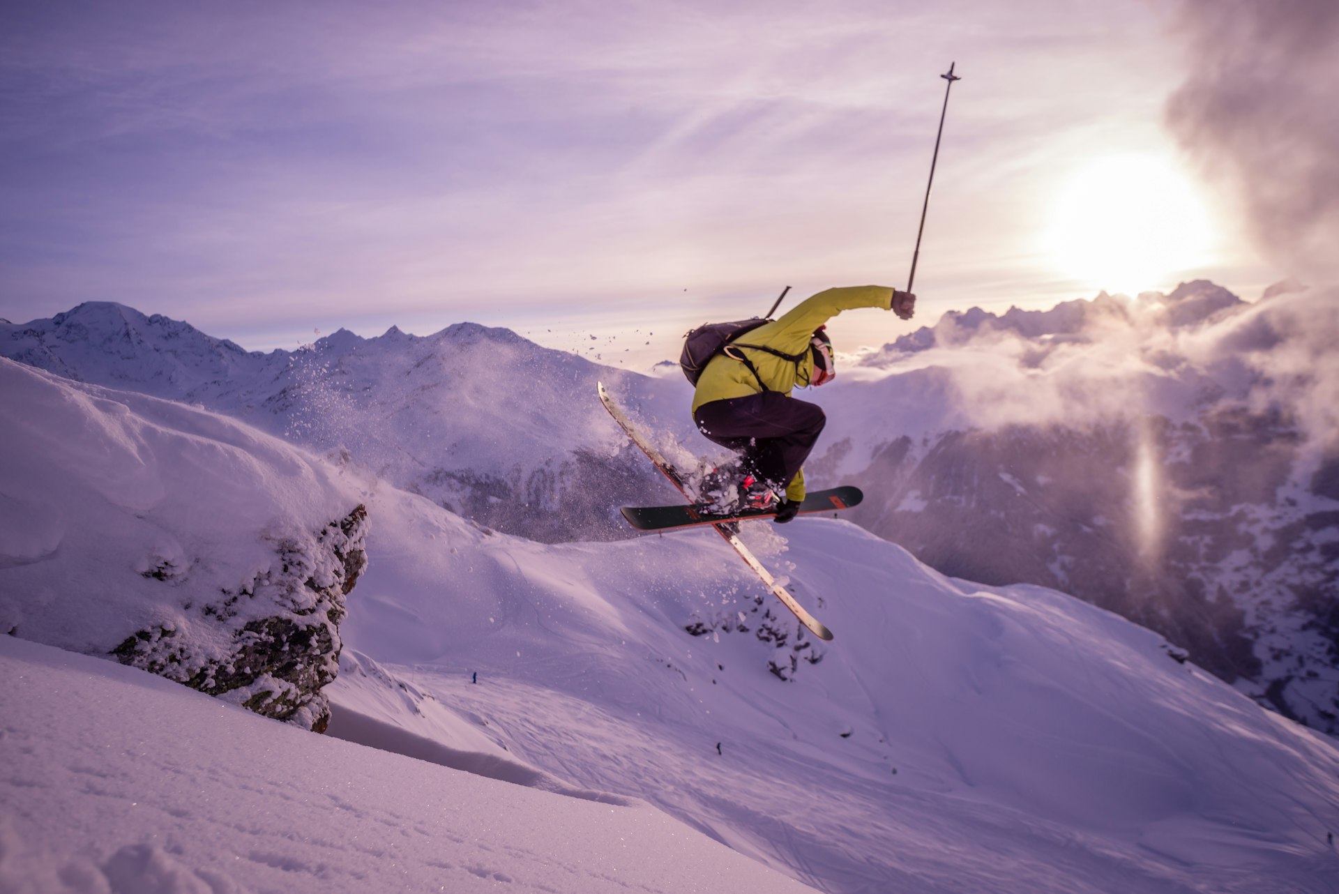 A skier flies through the air on the ski slopes above Verbier in the Swiss Alps. The mountains are blanketed in fresh snow and the sun is setting behind the mountains.