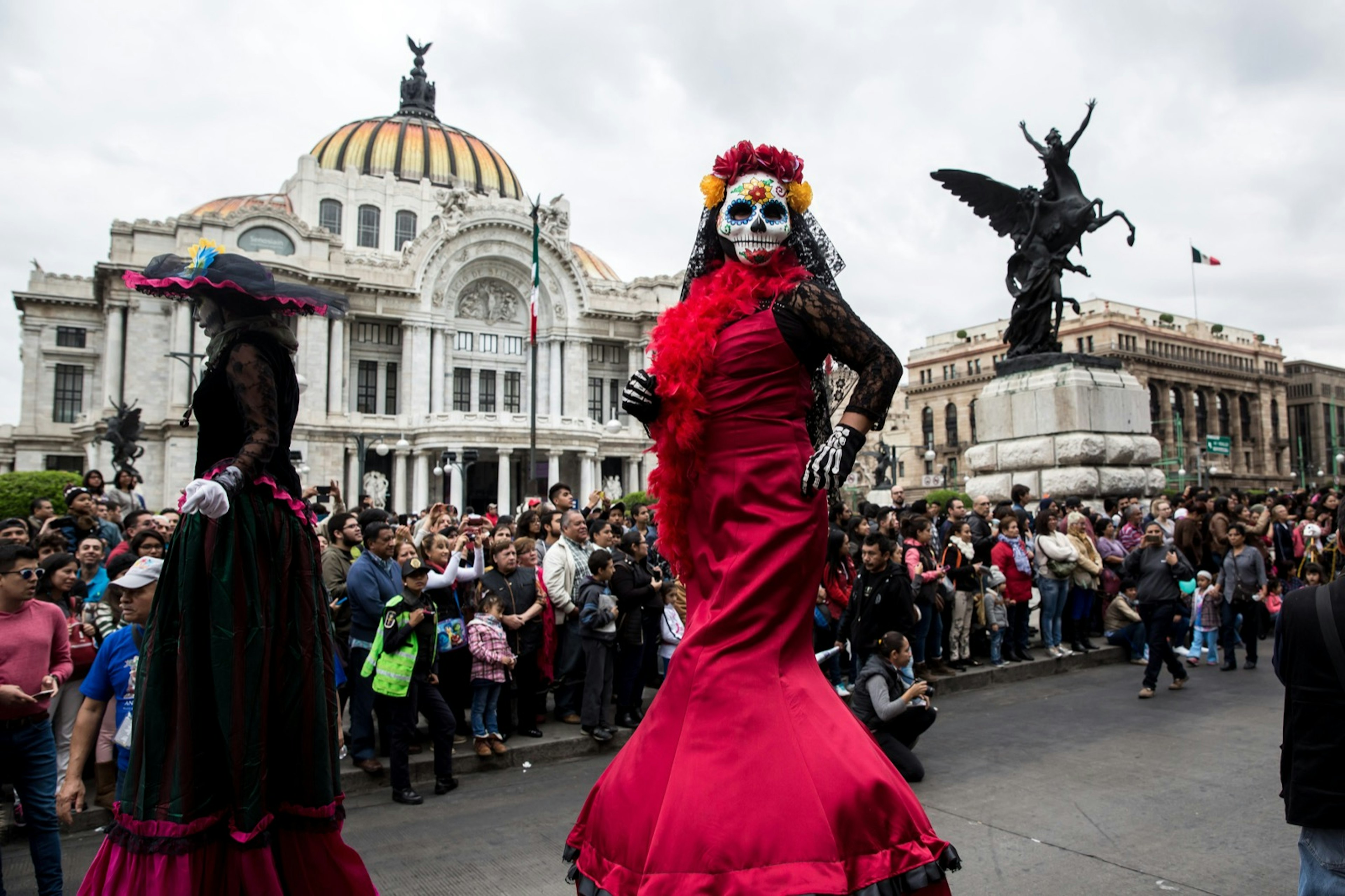 Performers in opulent clothing fringed with lace and skull masks walk down a road during a Dia de Muertos parade.