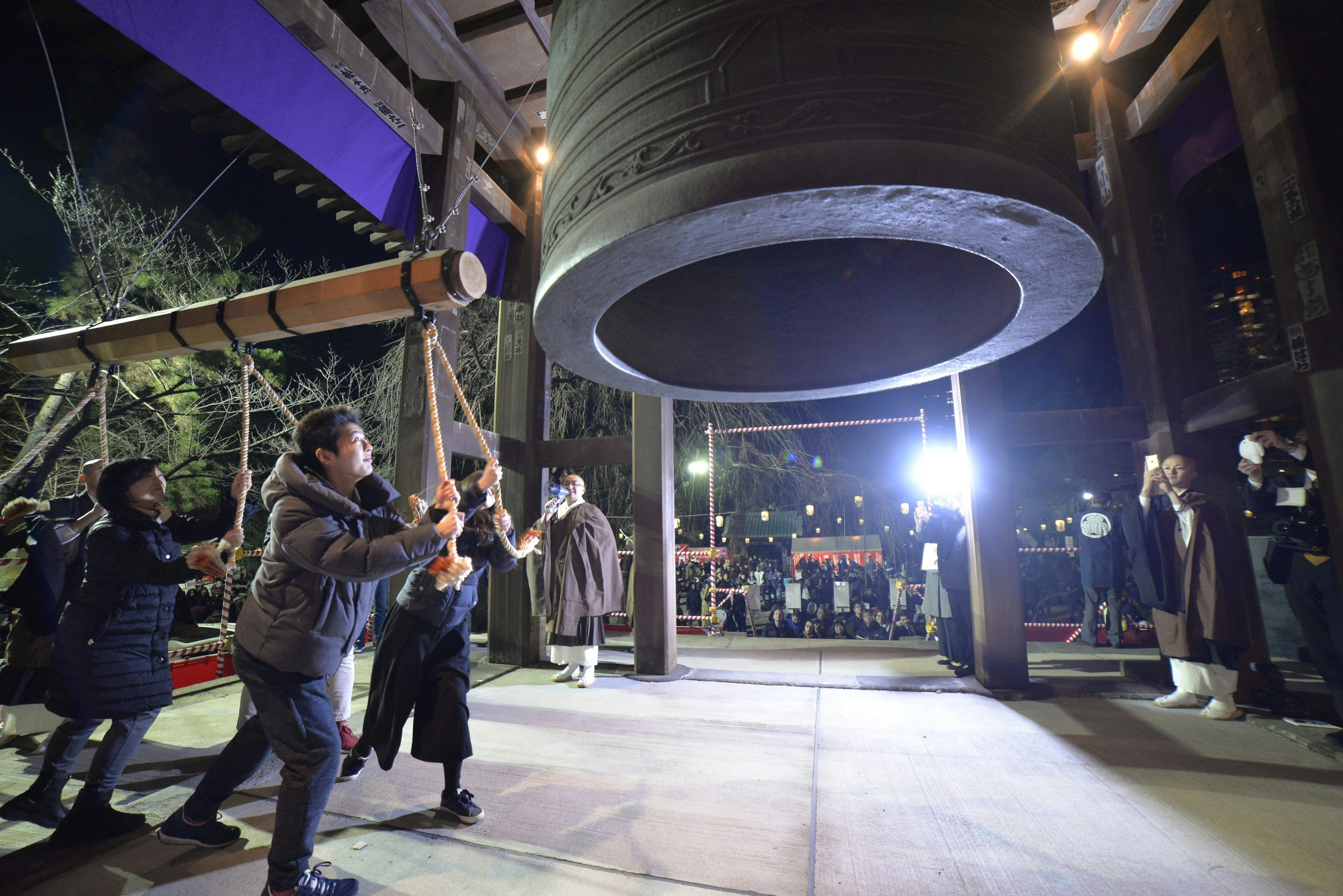 A group of people are holding ropes attached to a large wooden pole, which they are swinging to ring a huge bell in a temple. Monks in brown robes are watching and taking photos.