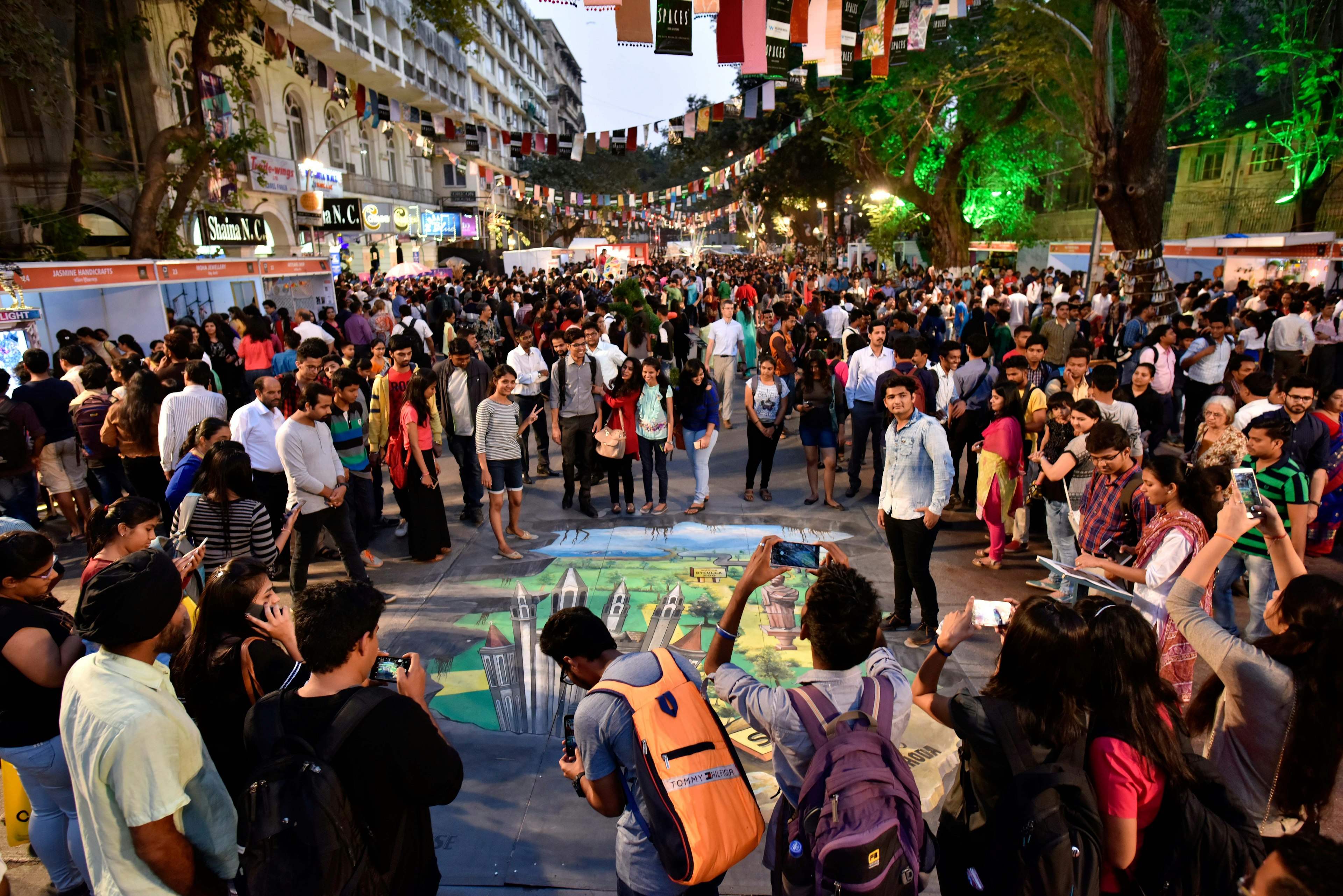 A huge crowd of people surround a large art mural drawn on the floor of Kala Ghoda in South Mumbai. Overhead colourful flags crisscross the street, which has other art displays in the distance.