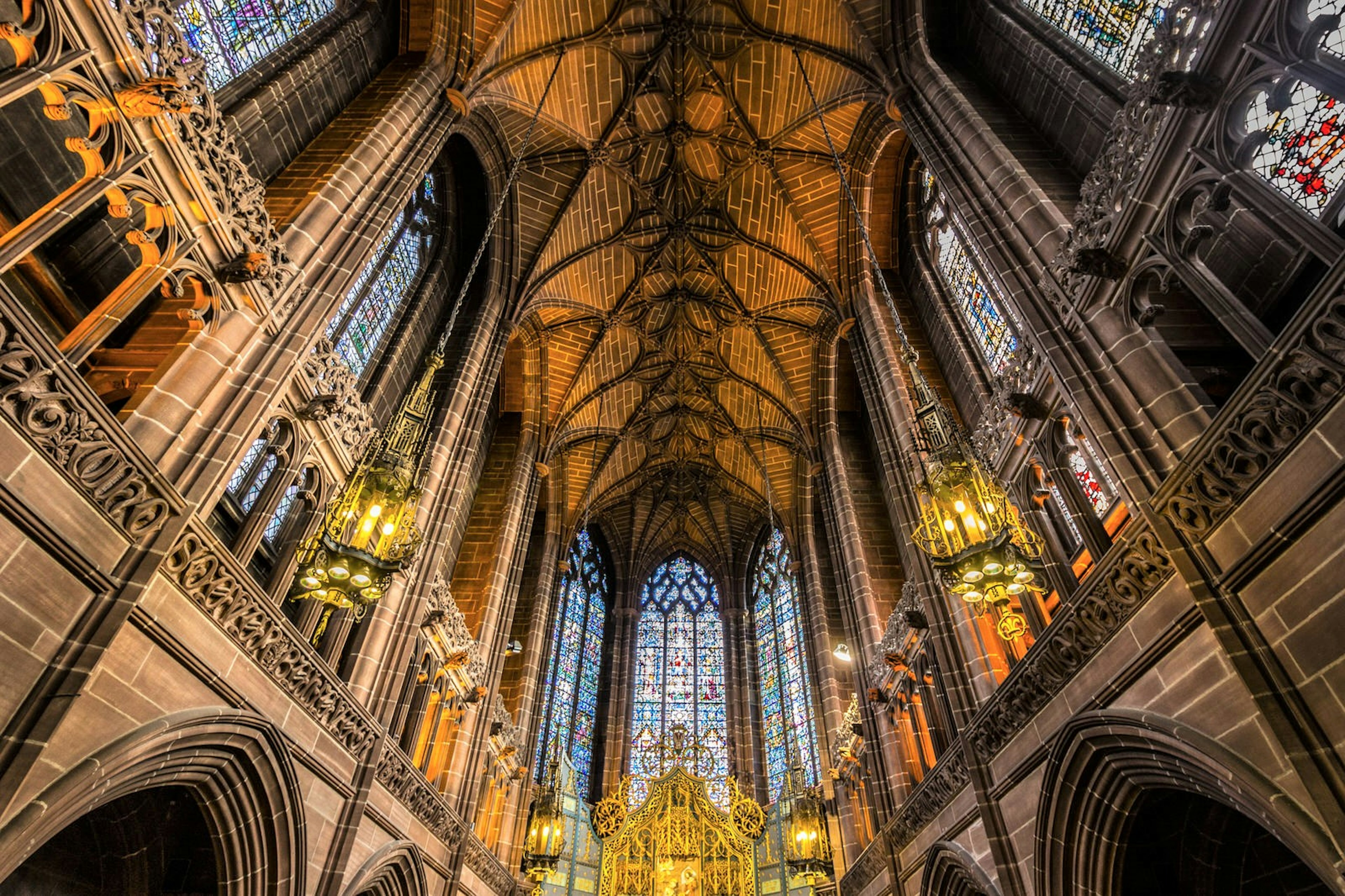 Liverpool's Anglican Cathedral's gothic roof is lit by yellow-hued lamps. The colourful stained-glass windows are a dramatic feature.