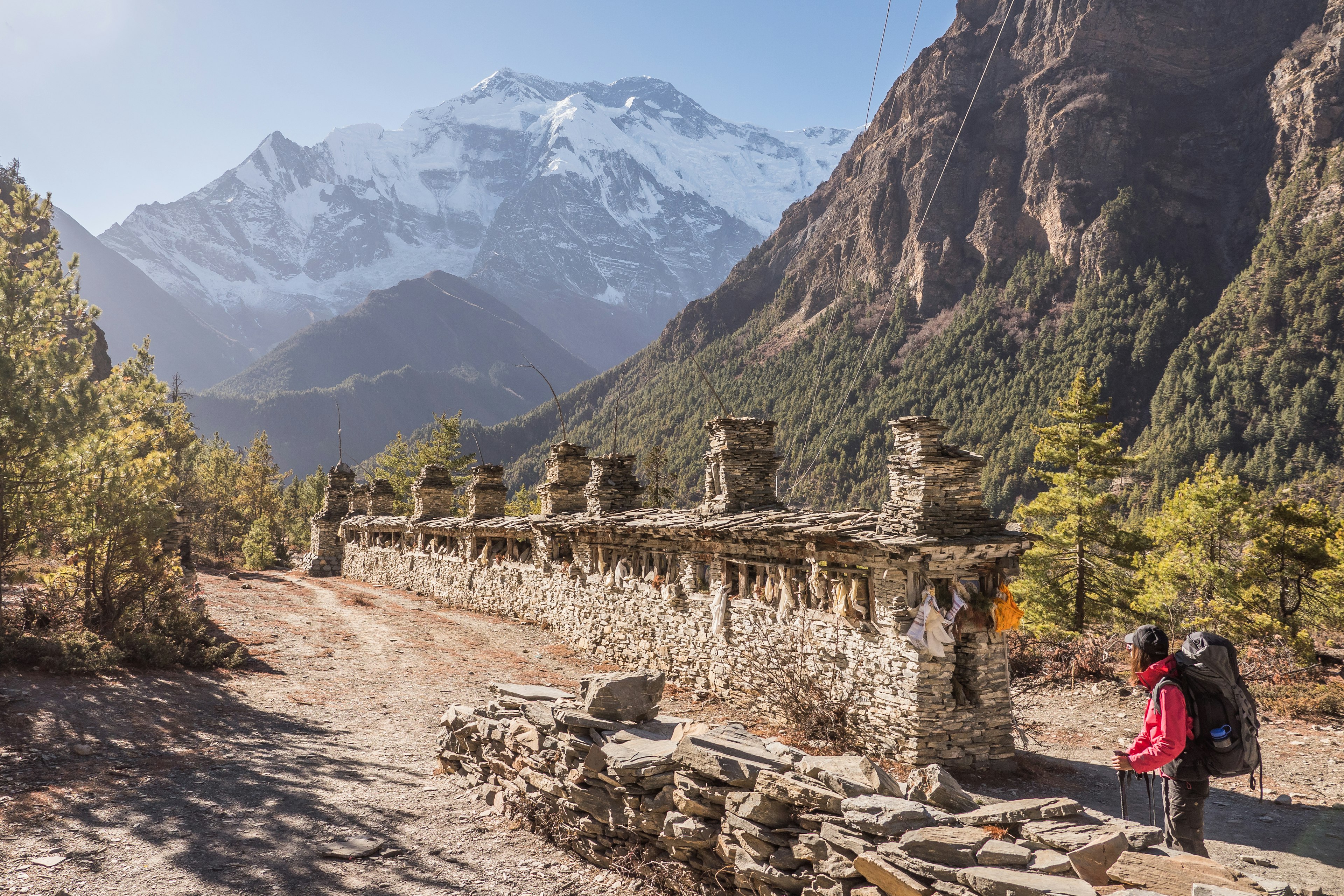 A female hiker in dark hiking pants and a pink long-sleeved shirt, dark-colored backpack stands in front of a long rock wall with crenellations and small chimney extends towards a huge massif