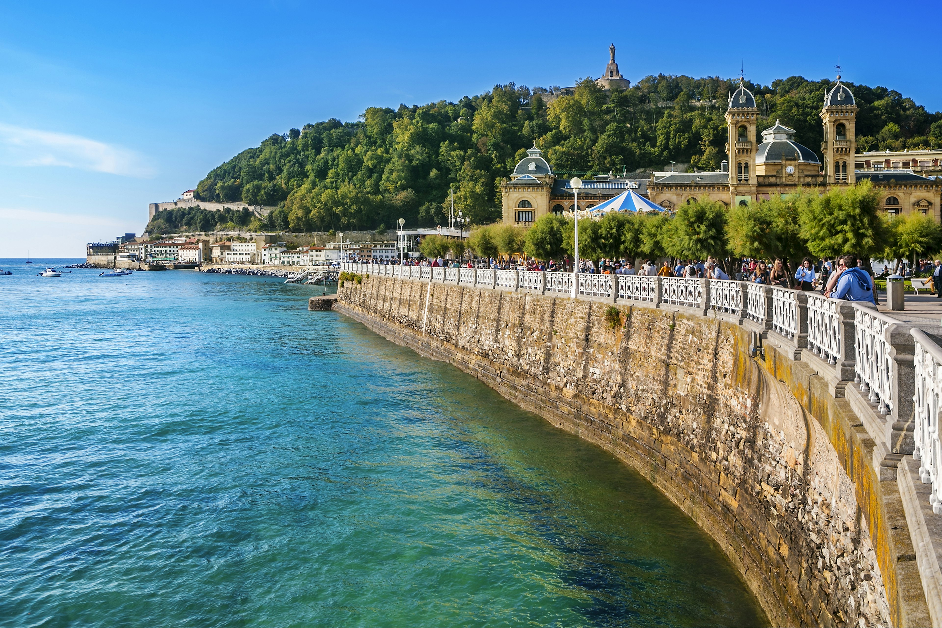 A group of people walk across a bridge in San Sebastián. There's a towering structure and large trees in the background; visit Basque country