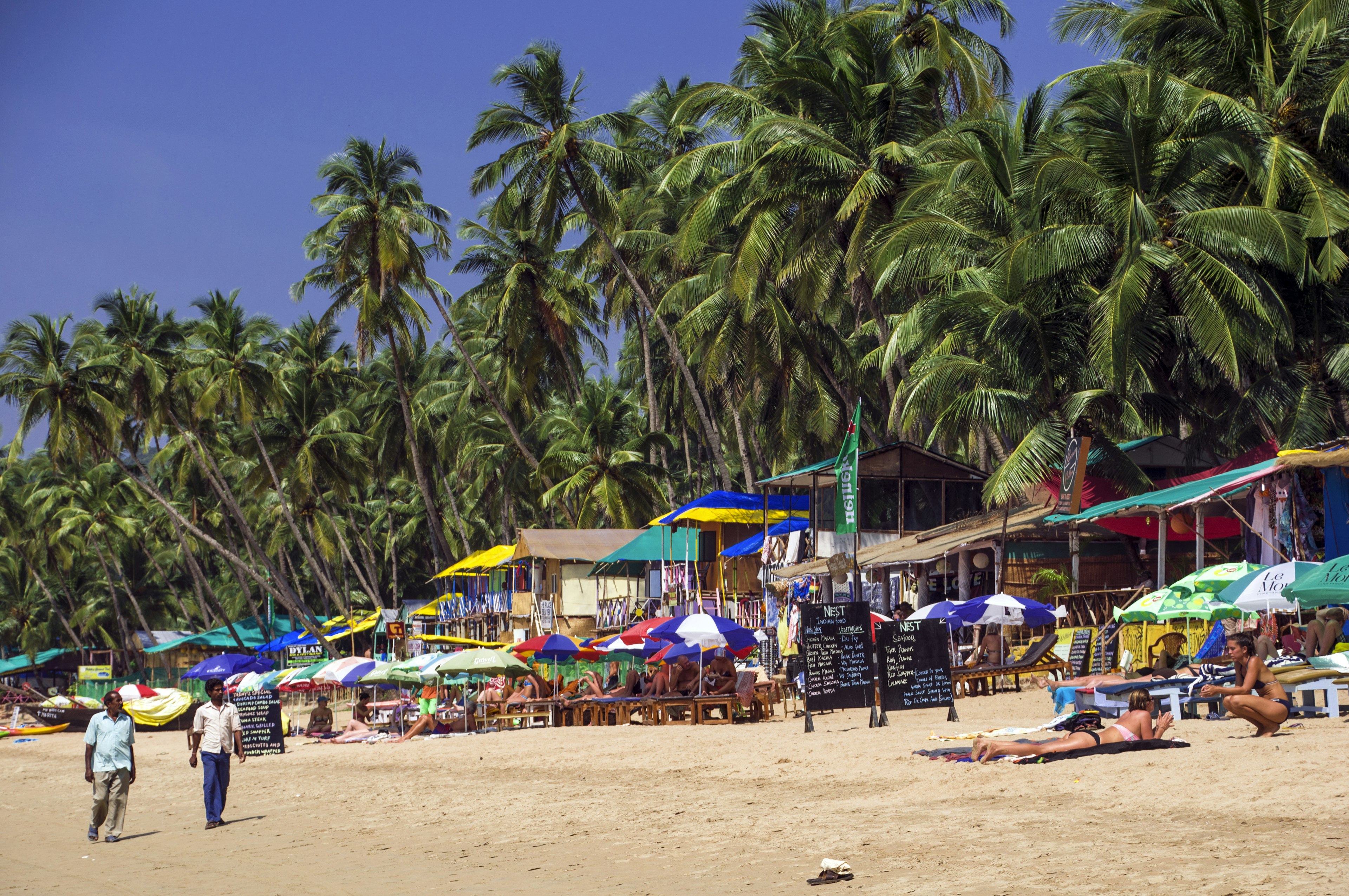 Two Indian men, one in khaki pants a light blue shirt, the other in dark blue pants and a light button-down, stroll along Palolem Beach in Goa past colorful umbrellas and sunbathing tourists. Behind the row of umbrellas and beach huts with bright awnings are a thick grove of palm trees.