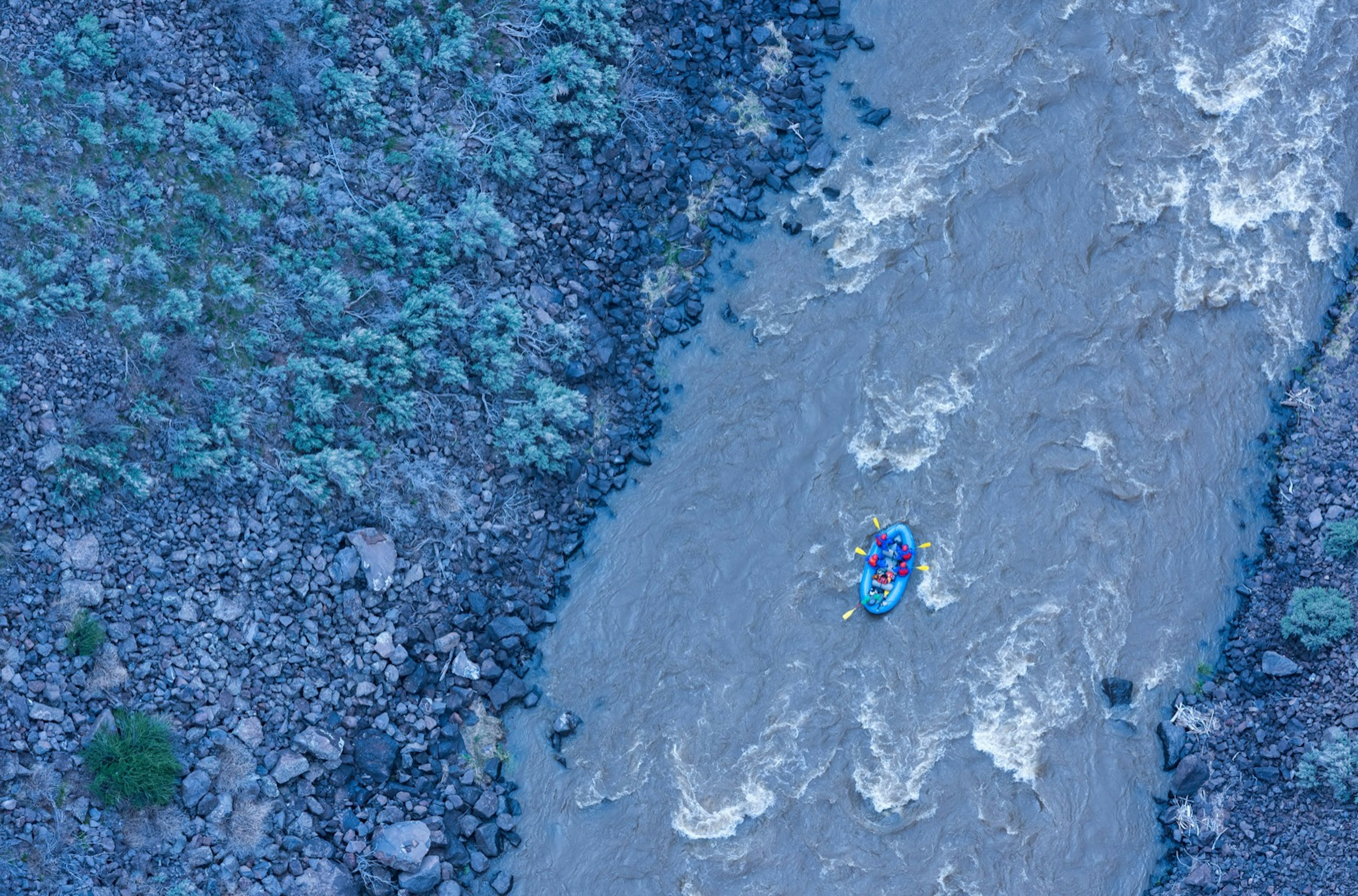 Looking straight down on the Rio Grande river from very high at twilight, as a single whitewater raft navigates some rapids.