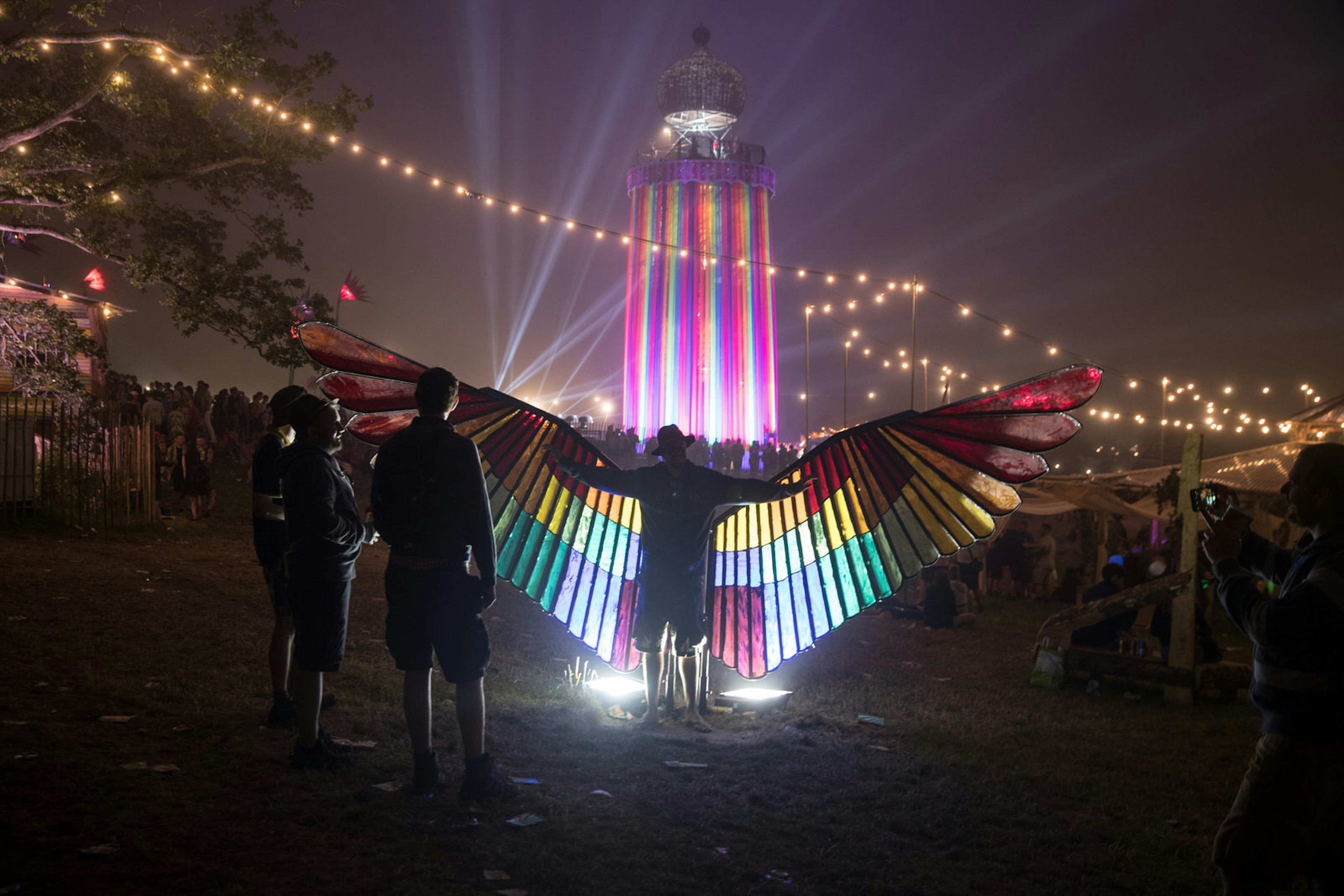 Someone wearing huge rainbow coloured wings that are lit up by a colourful ride in the background