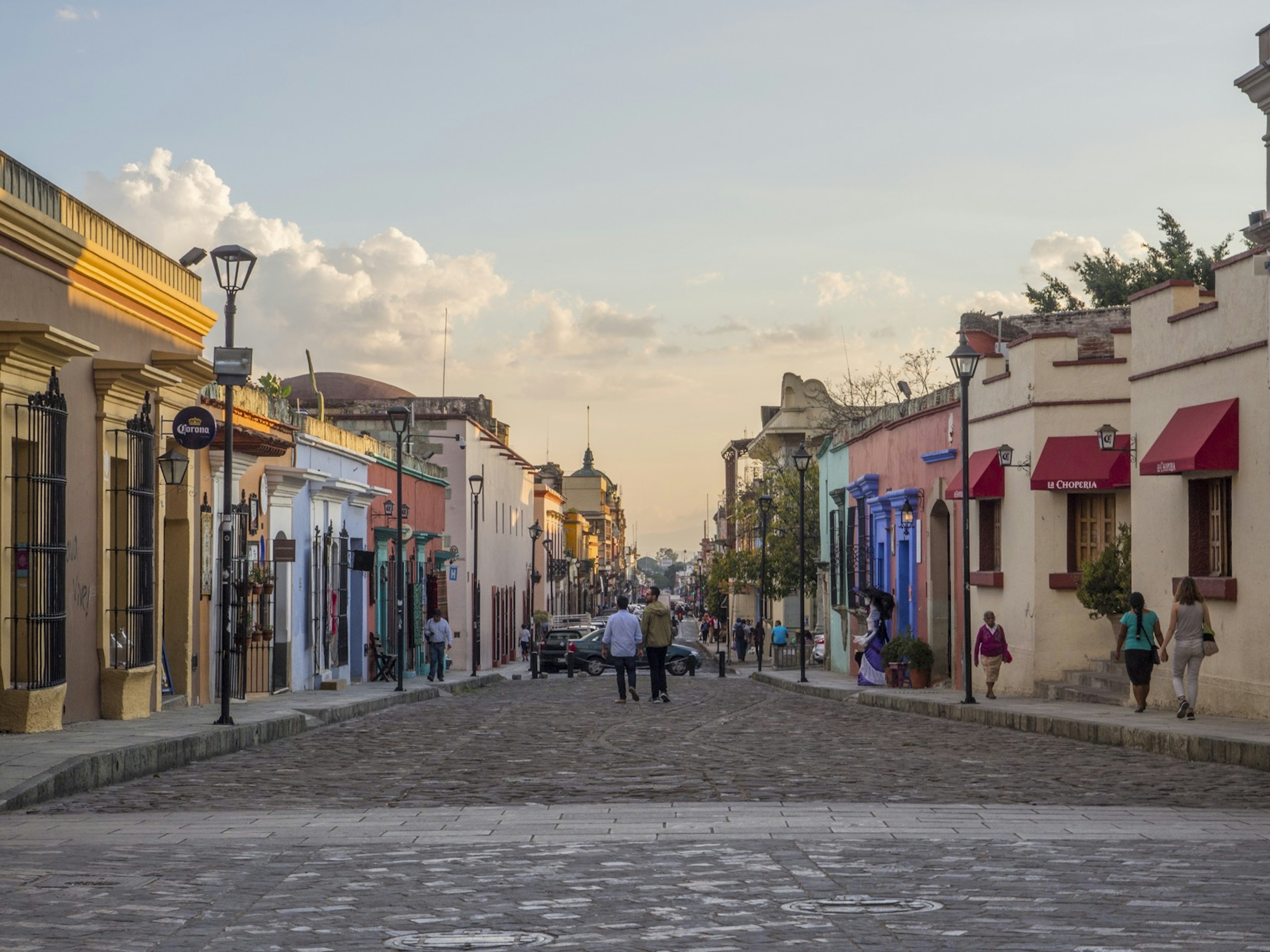 Two men stand in the middle of a cobble stone street lined with colorful buildings and street lamps some people walk on the sidewalks the sun is setting casting a golden glow in LGBTQ-friendly Oaxaca