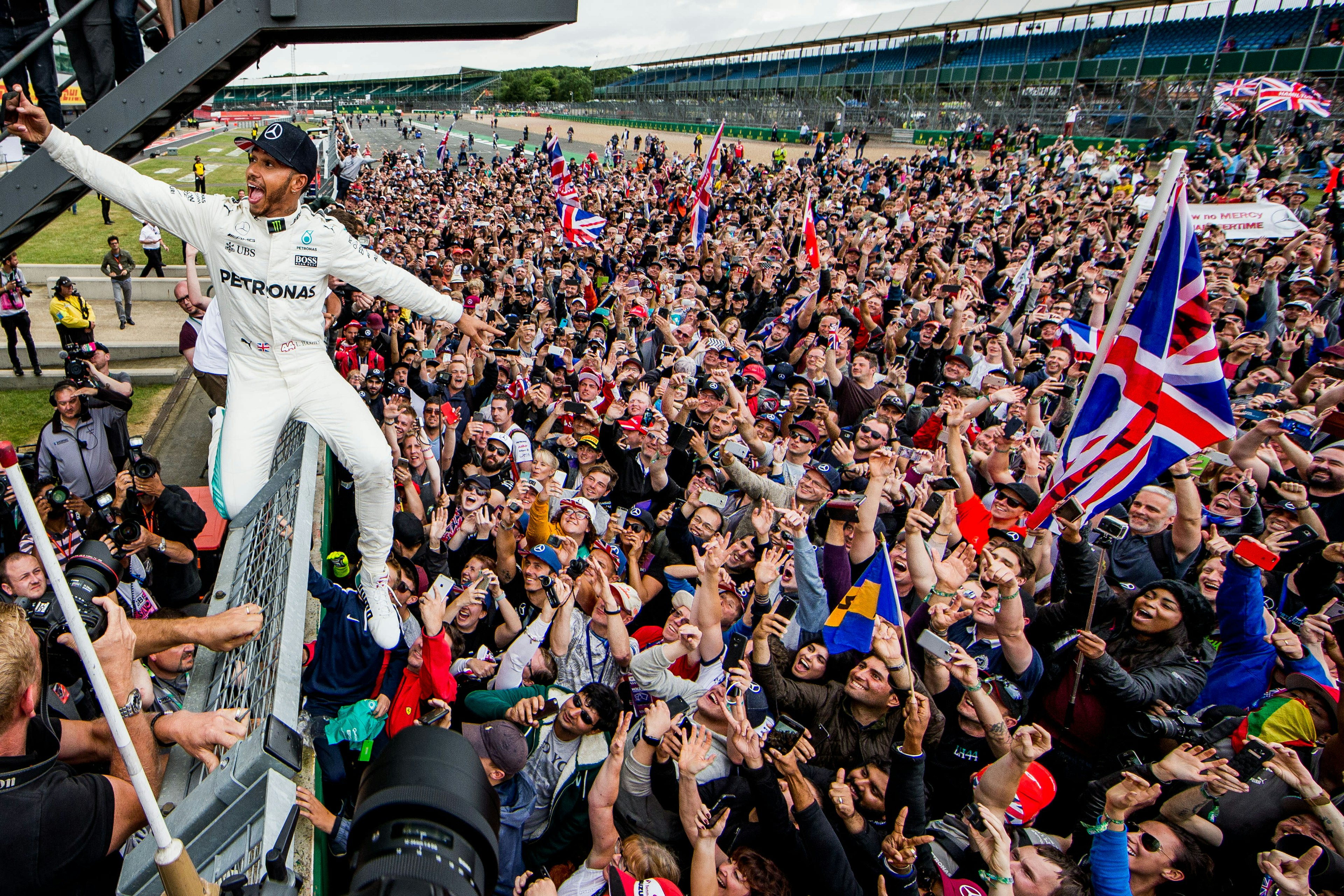 Legendary British Formula 1 driver Lewis Hamilton celebrates with the crowd at Silverstone in 2017. Lewis sits on top of a high fence and takes a selfie with the crowd in the background. They are all waving at him and waving the British flag.