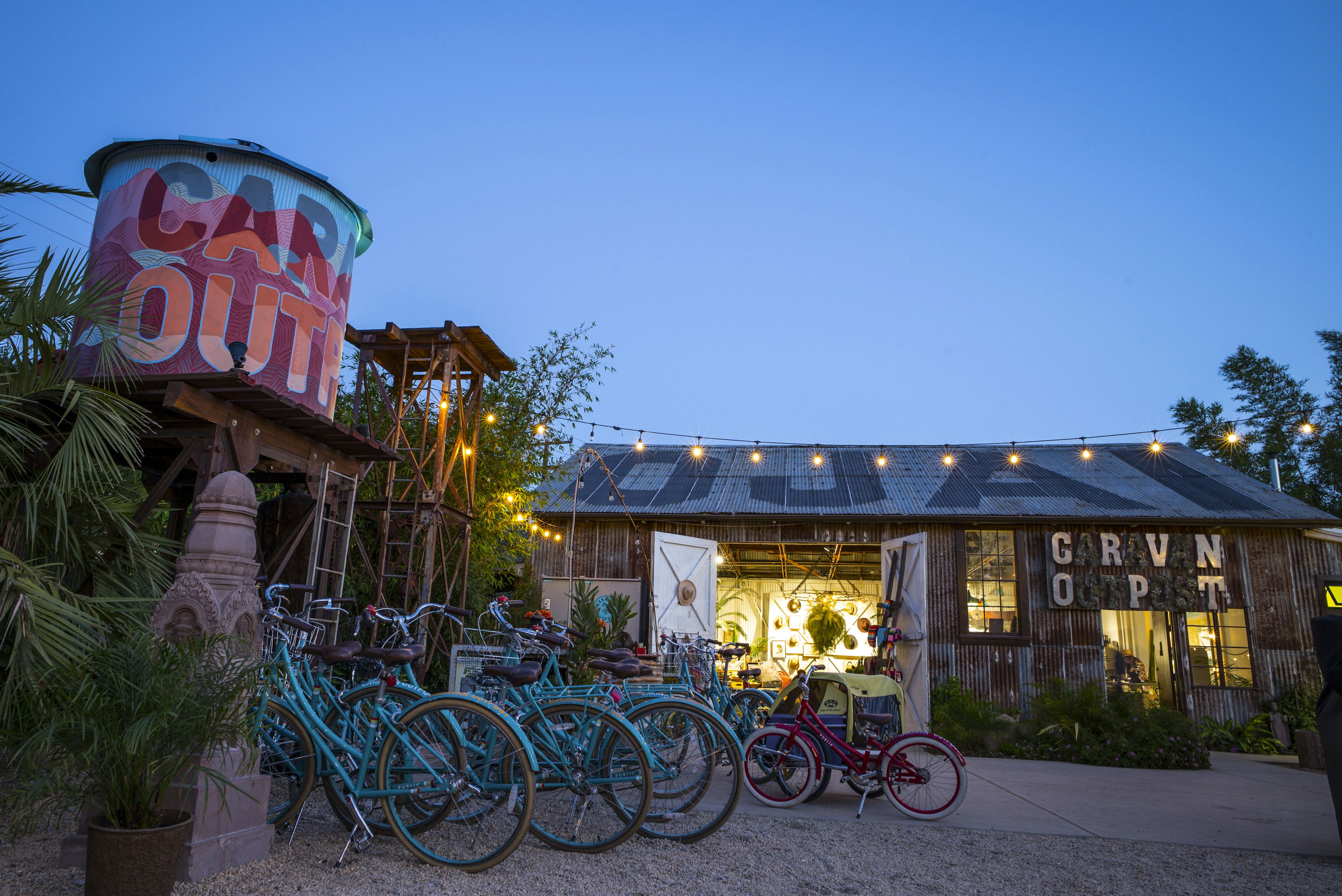 Against a bright blue evening sky sits the tin roof of Caravan Outpost, painted to read OJAI in large black letters. Two white doors are flung wide open, revealing the yellow glow of the lights inside. Overhead is a yellow glowing row of string lights. A water tower on the left side of the frame paitned in pinks and oranges to mirror the surrounding mountains reads Caravan Outpost. Several bright blue bikes and one kid-size magenta bike sit on a gravel lot near a cluster of palms