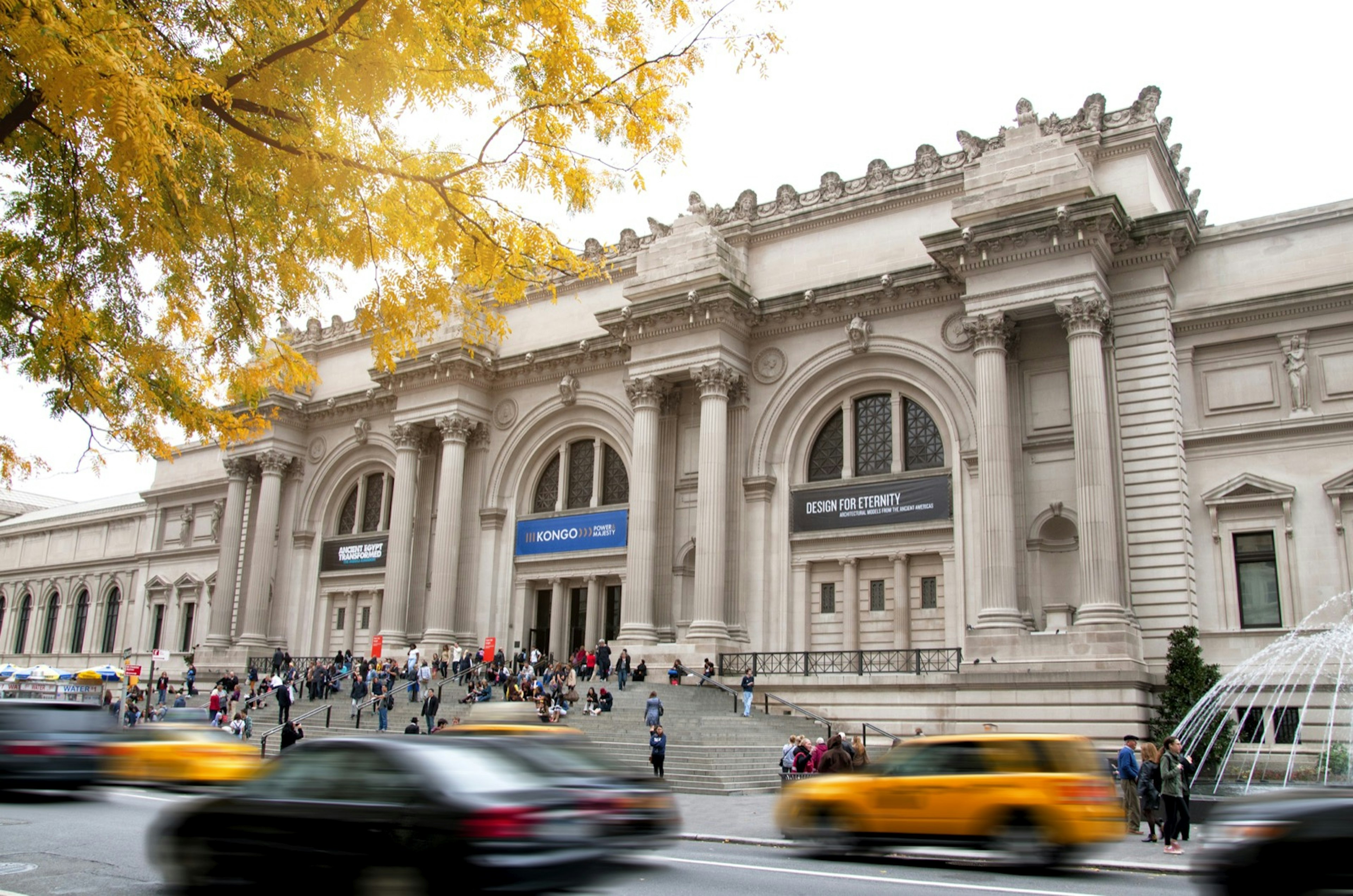 Cars and taxis pass by the facade of a museum as people congregate on the steps