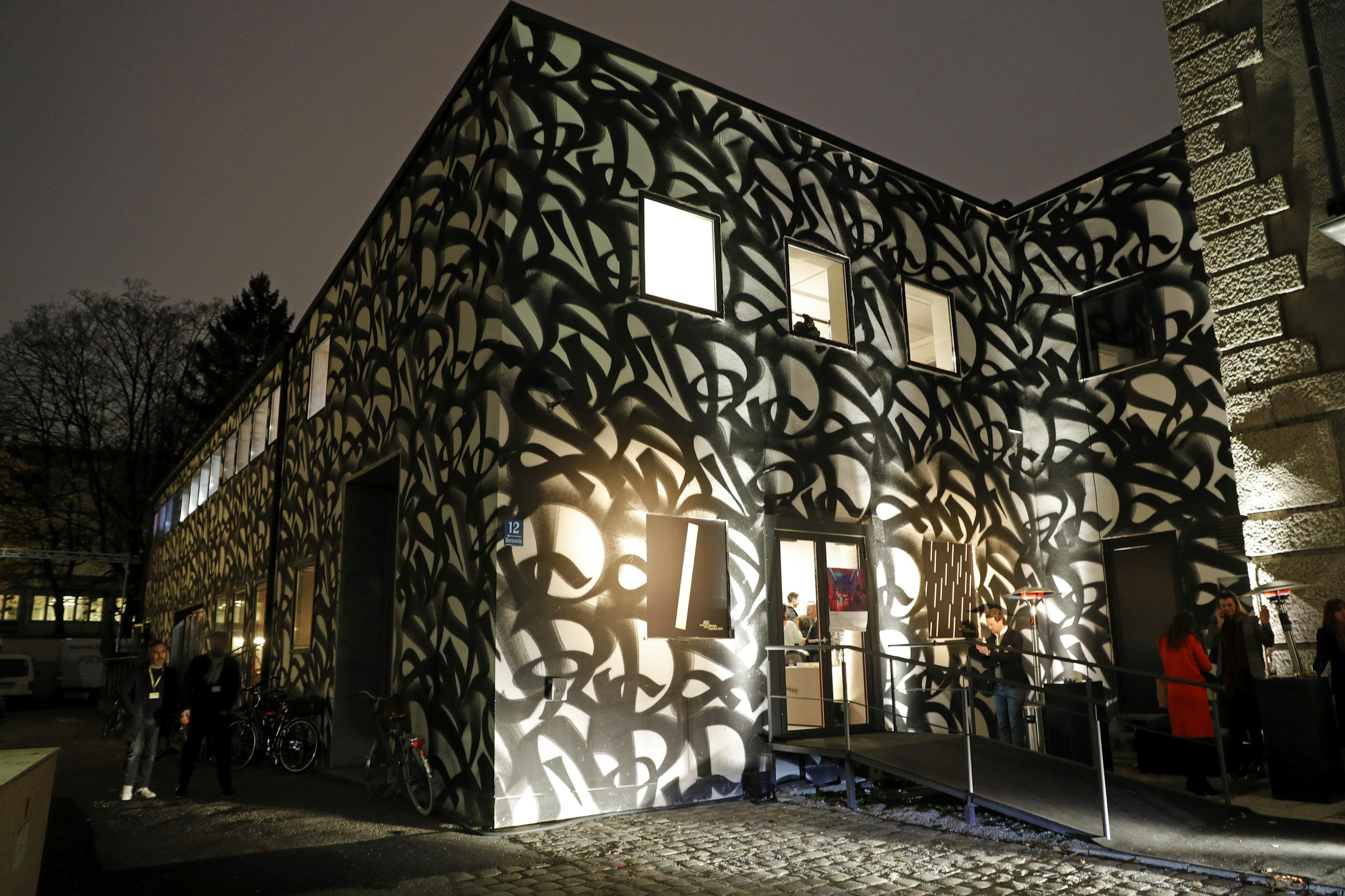 Nighttime exterior shot of an art gallery. The walls are entirely covered in black abstract graffiti against a white background. Some people are milling around outside.