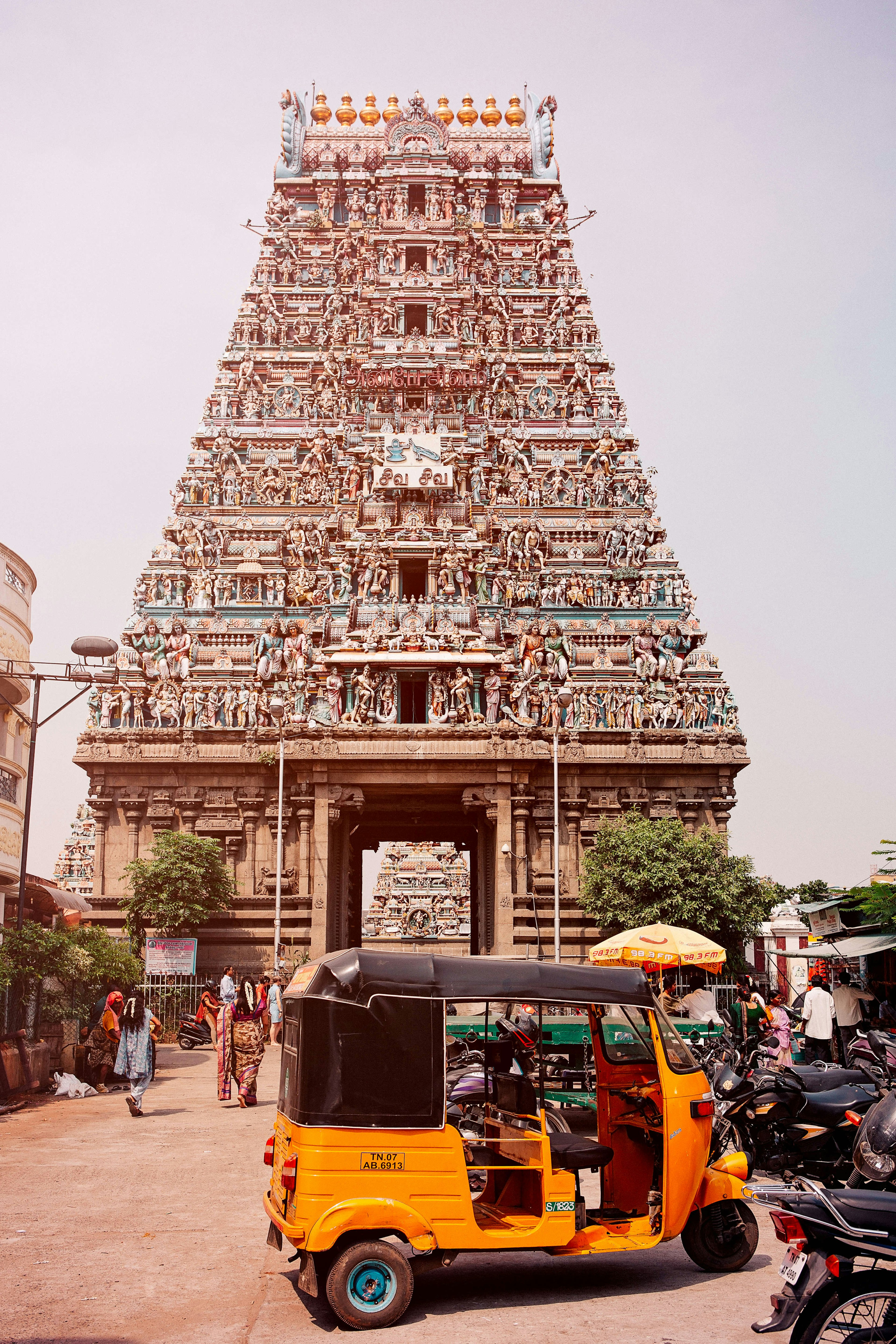 A bright orange motorized rickshaw with a black roof is parked in front of the colorful facade of the the Kapaleeswarar Temple in Chennai, which has several tiers like a pyramid covered in sculptures of humans and animals.