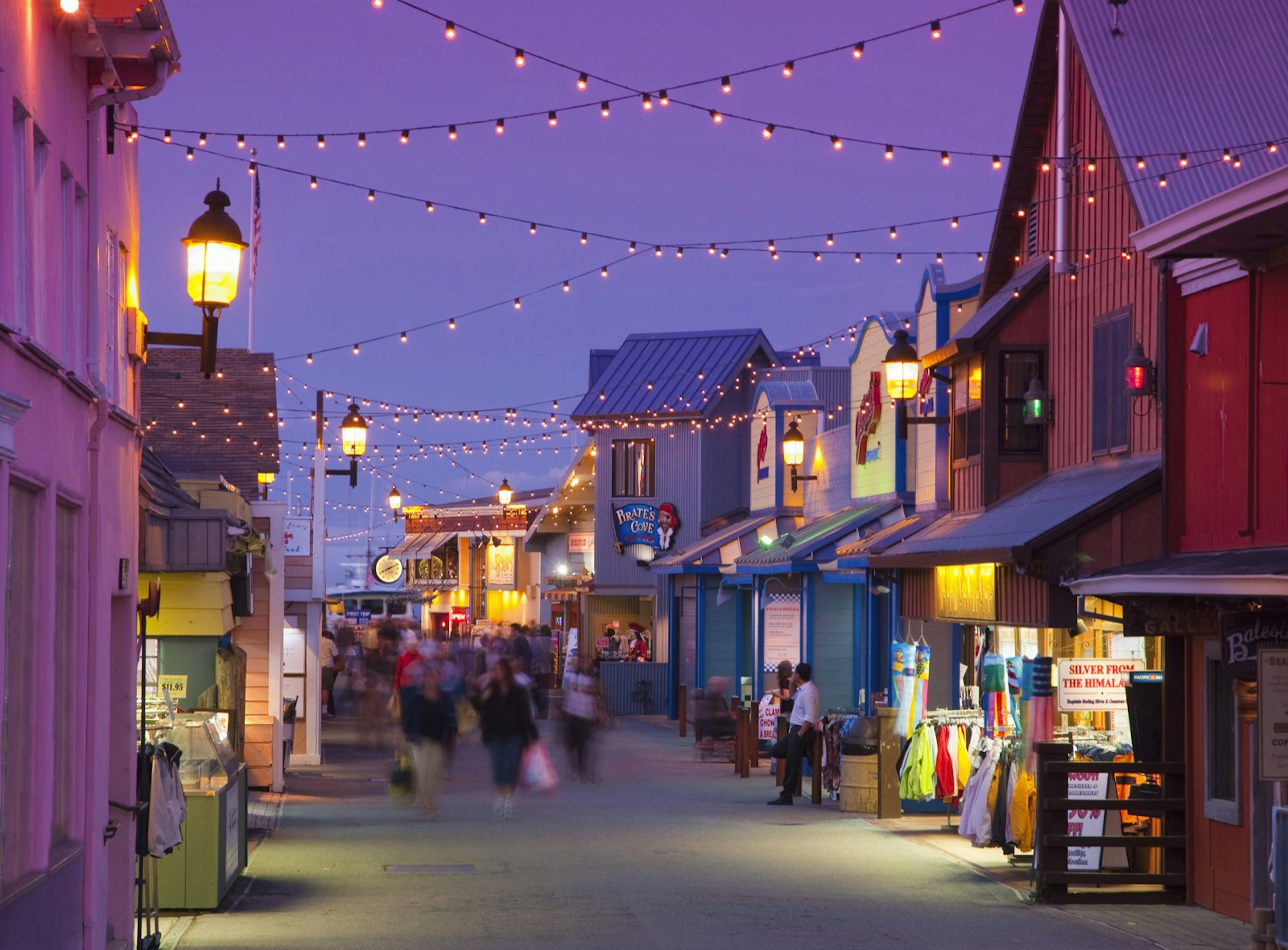 Twinkle lights sparkle above a street lined with wooden structures and people walking in Monterey, California