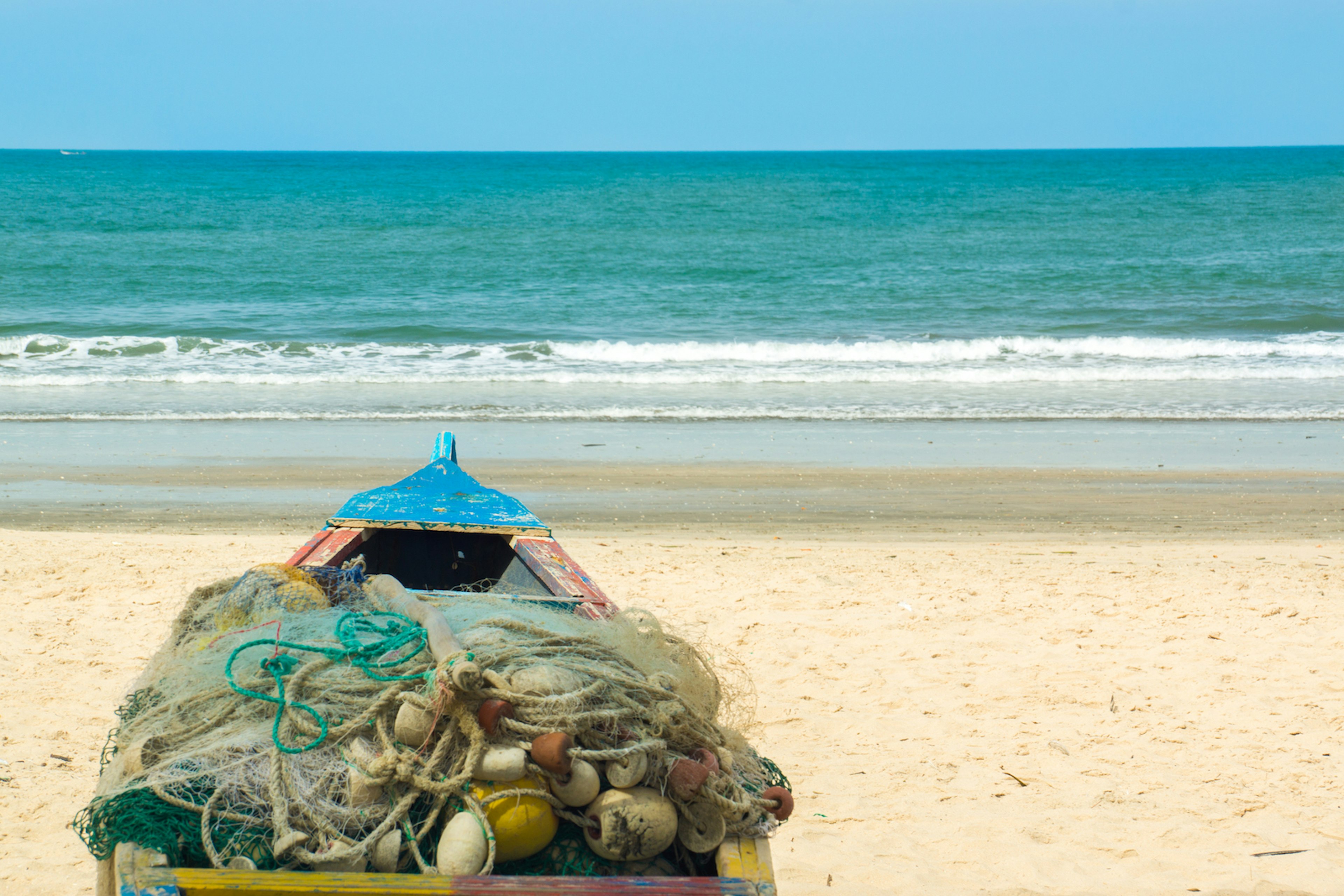 A colorful pirogue with a fishing net thrown on top on the beach and a view of blue-green water in Kotu