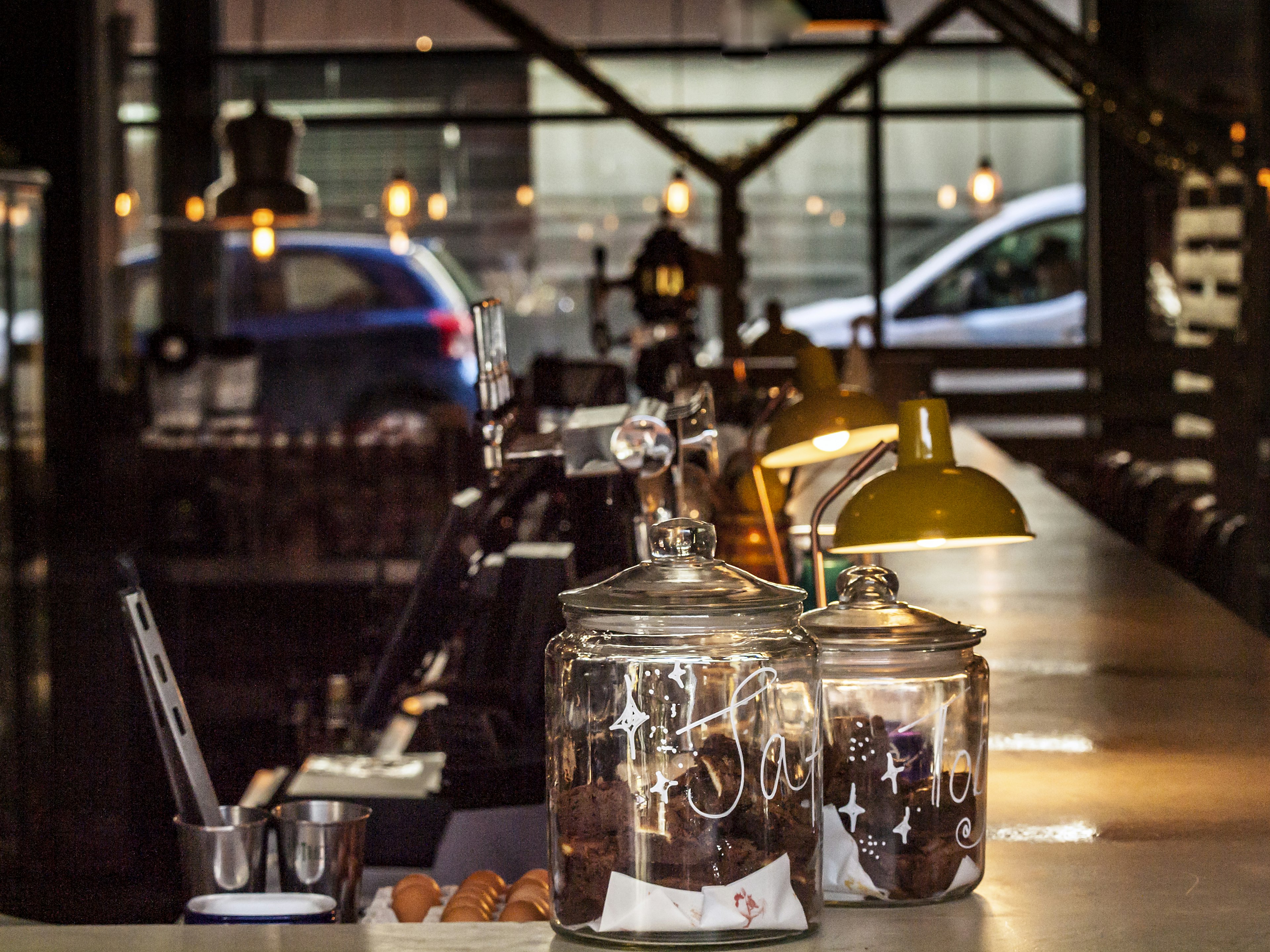 A warmly lit bar in a coffee shop, with utensils in pots and two large glass jars containing chocolate brownies.