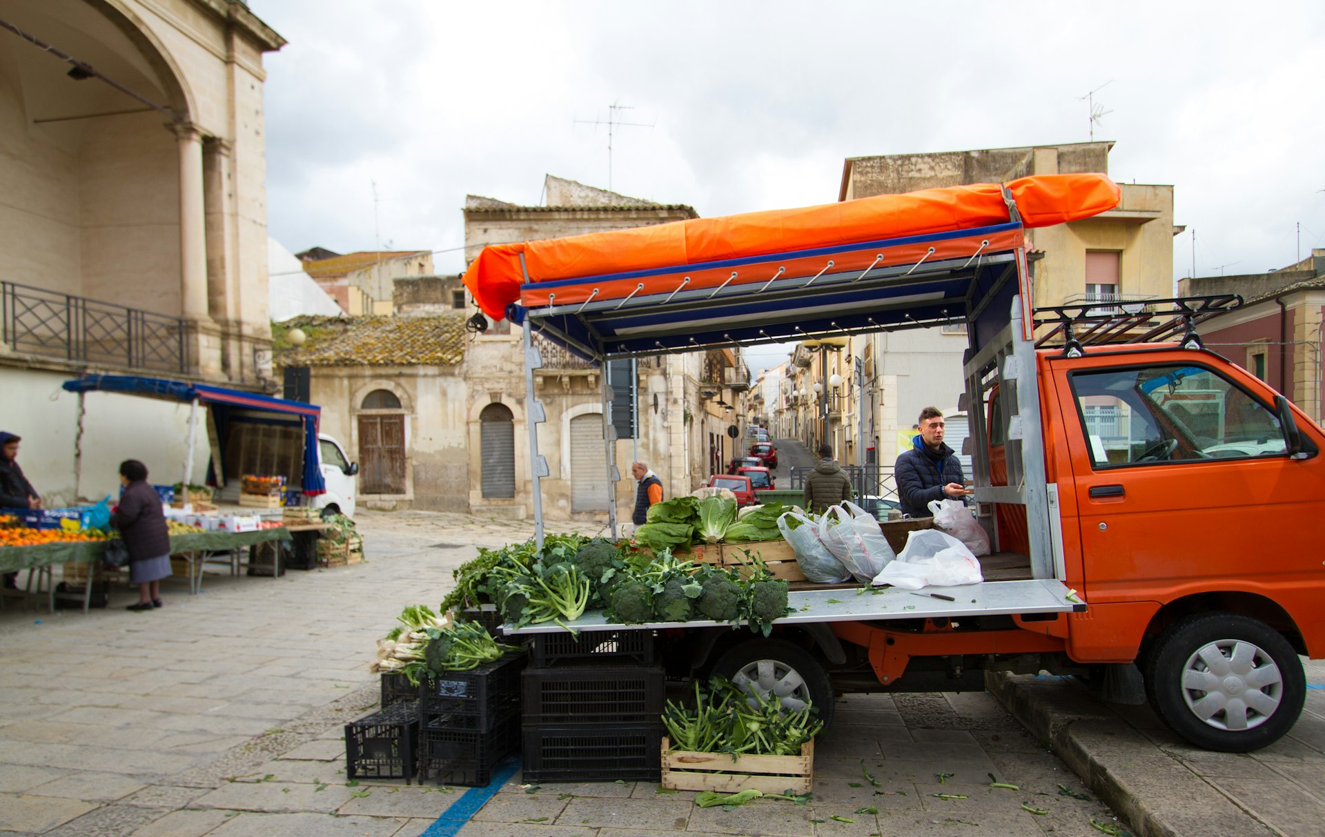 Fruit and vegetable vendors and one senior customer at an open-air farmer’s market in central Comiso, Sicily, in winter