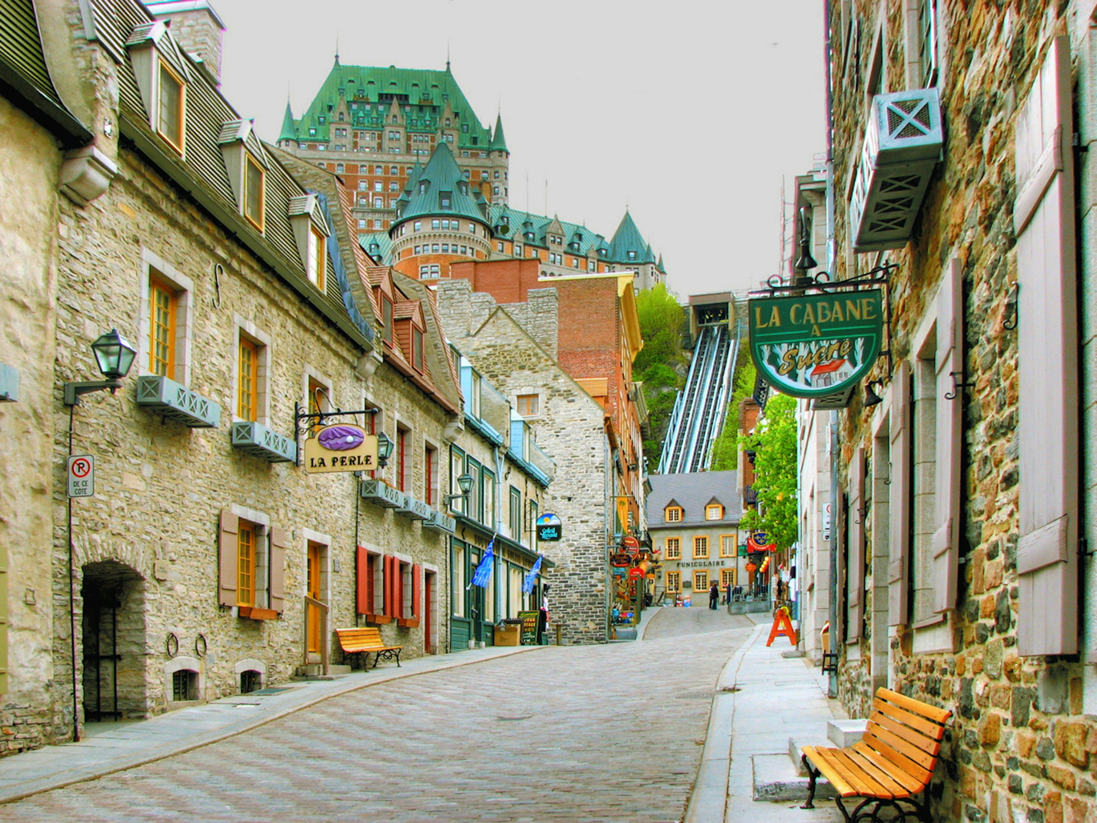 ϳé City’s cobblestone streets with stone buildings either side that have been turned into various shops and cafes. Château Frontenac, a large red brick building with a copper roof and turrets, is visible in the background.