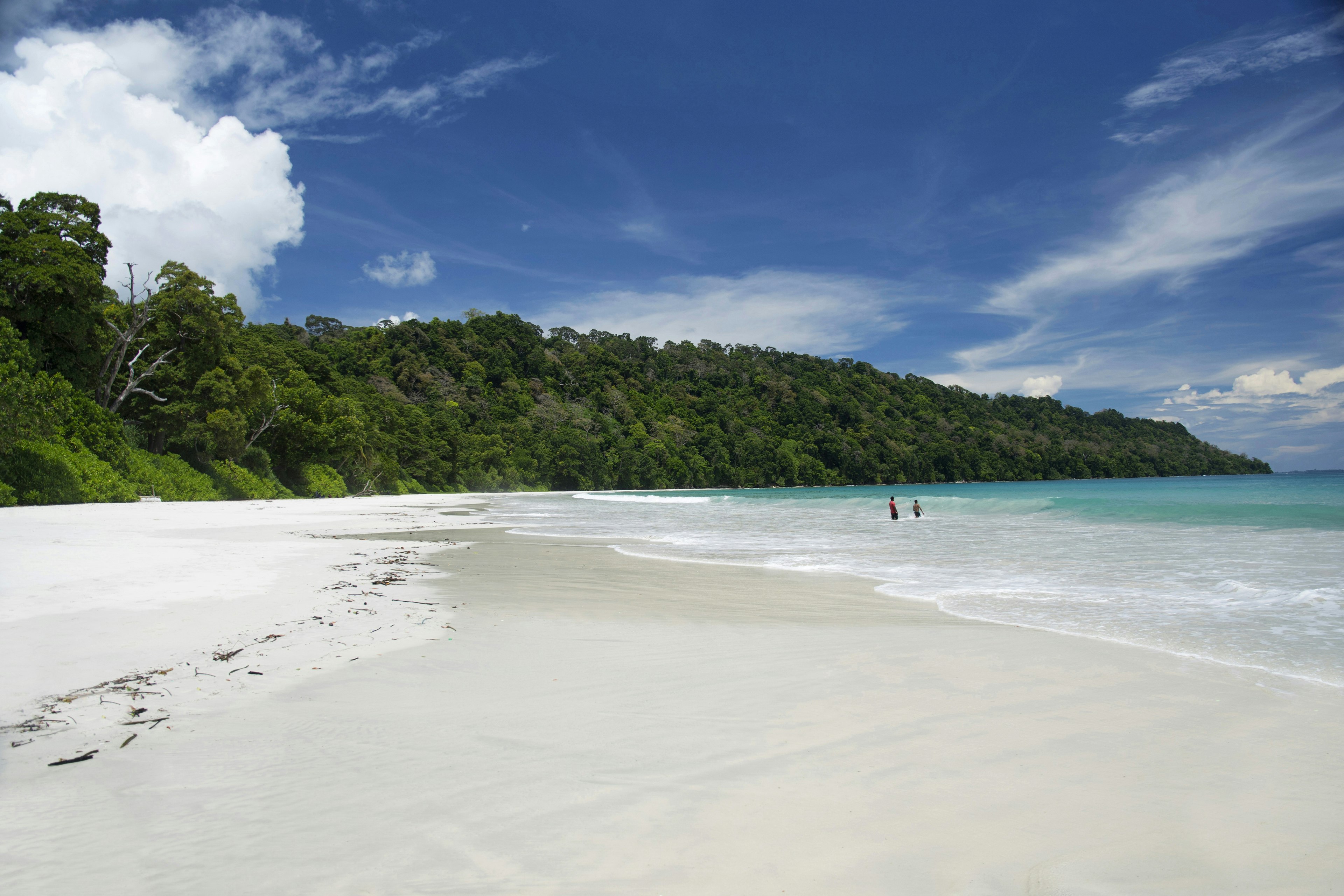 Two men paddle in the shallows of an otherwise empty beach on Havelock Island, which is part of the Andaman Islands archipelago. The white-sand beach is backed by forest.