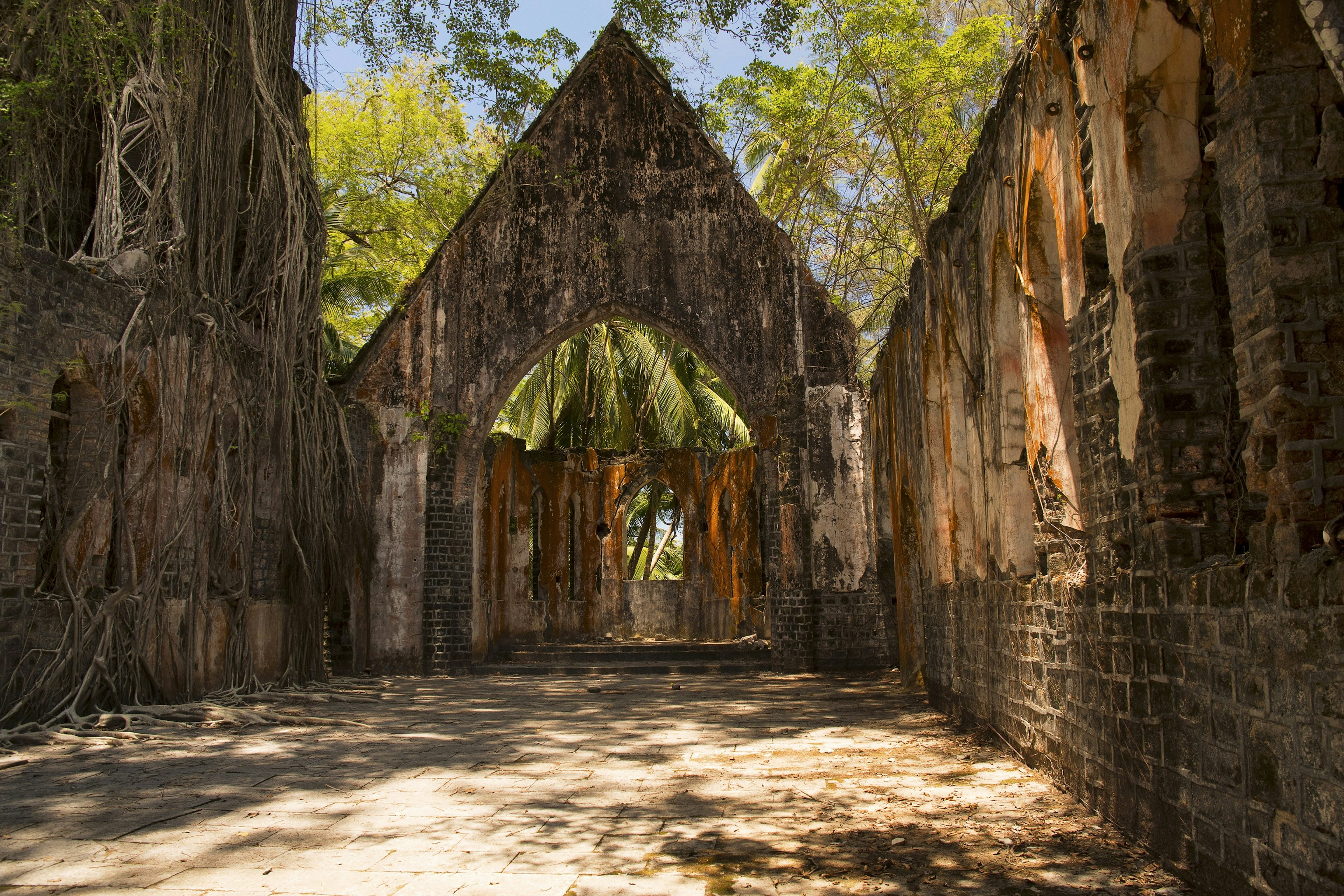 The shell of an old Protestant church built of stone masonry, with windows made from Burma teak, that still stands on Ross Island in the Andamans. The church is missing its roof, and through the hole - and window gaps - the green jungle is visible.