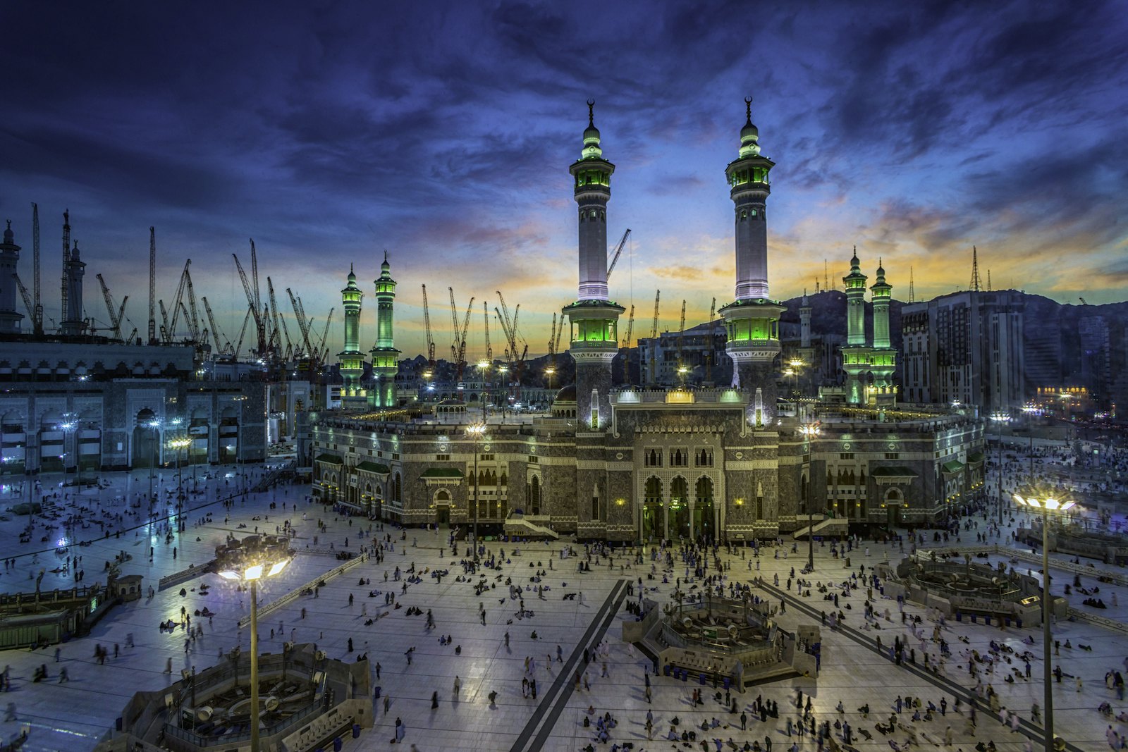 Large groups of people are making their way towards the the mosque on all sides. The sun is setting and the scene is lit up by the towers of the mosque glowing in the dusk. Hajj diaries