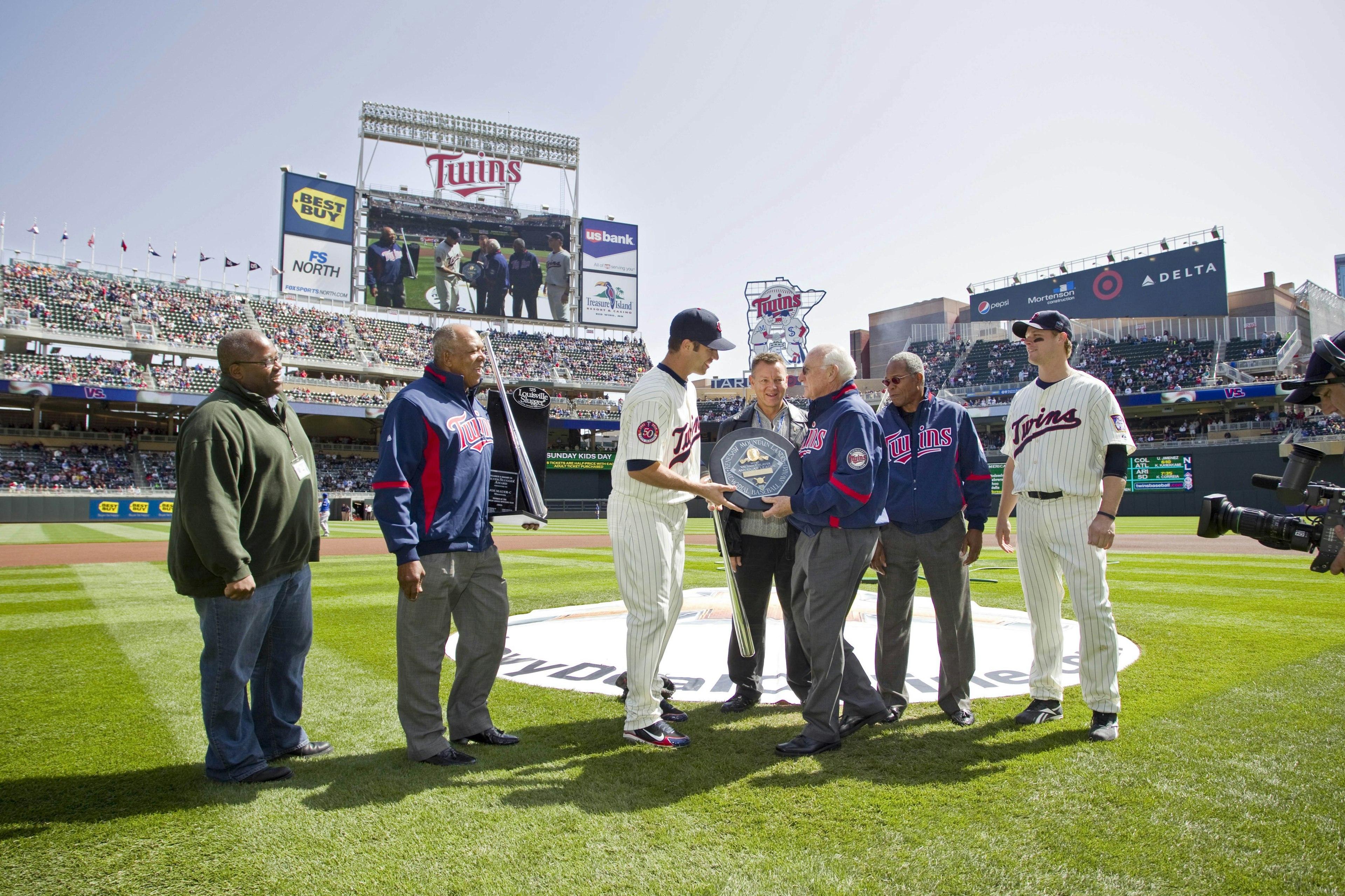 A shot of the Louisville Slugger Stadium from the field, where members of the Minnesota Twins in white striped uniforms and blue ballcaps are presented a trophy by former Twins players in blue letterman's jackets with red logos and grey slacks. Overlooking the presentation of the MVP plaque is a Louisville Slugger representative in a blue and red jacket with grey slacks and an African American gentleman in a green pullover and blue jeans.