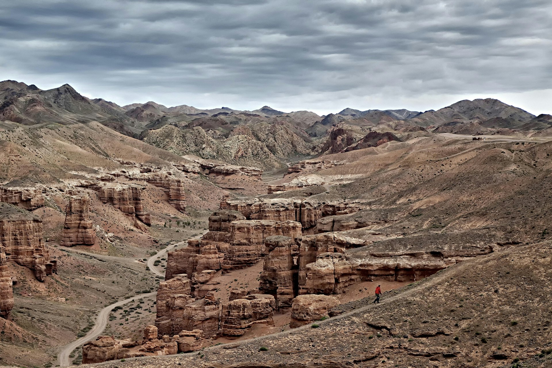 A sole hiker follows a trail alongside tall red rocky outcrops that fill a valley