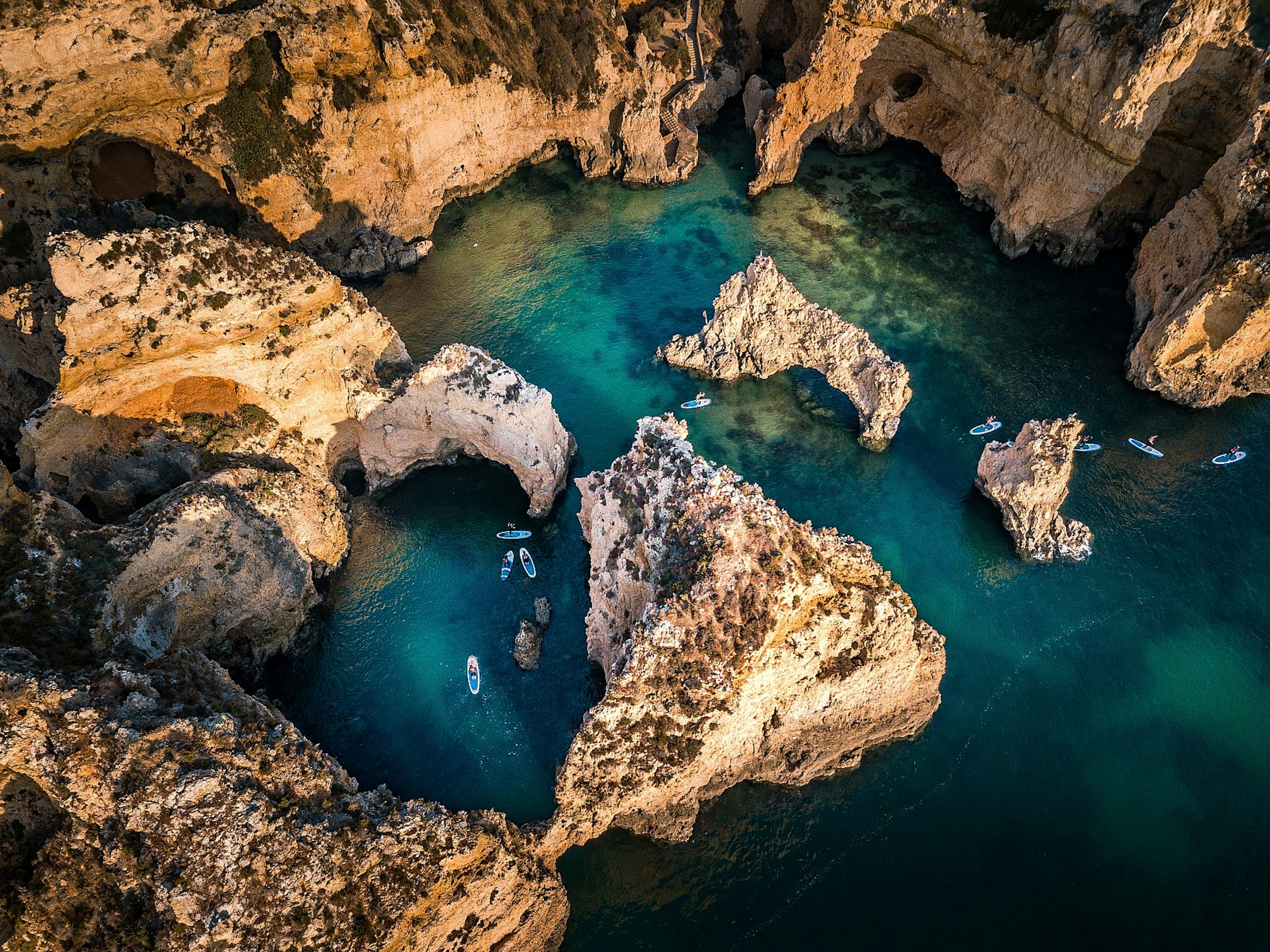 An aerial view over people stand-up paddle boarding amid golden sandstone cliffs and sea stacks, lapped by turquoise water.