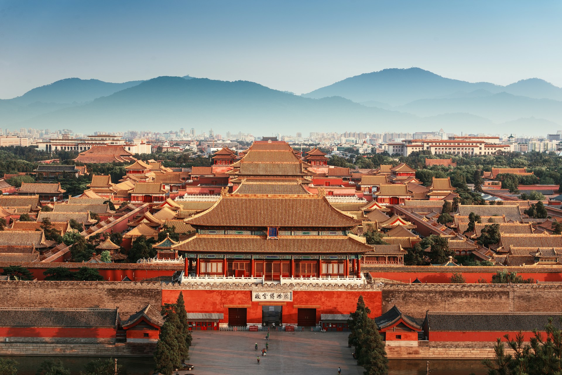 Aerial view of the Forbidden City in Beijing, China. The image shows the sprawling complex of traditional Chinese imperial buildings with golden roofs and red walls. In the foreground is the main entrance gate with a large courtyard. The city skyline and misty mountains are visible in the background, creating a striking contrast between ancient architecture and modern urban landscape.