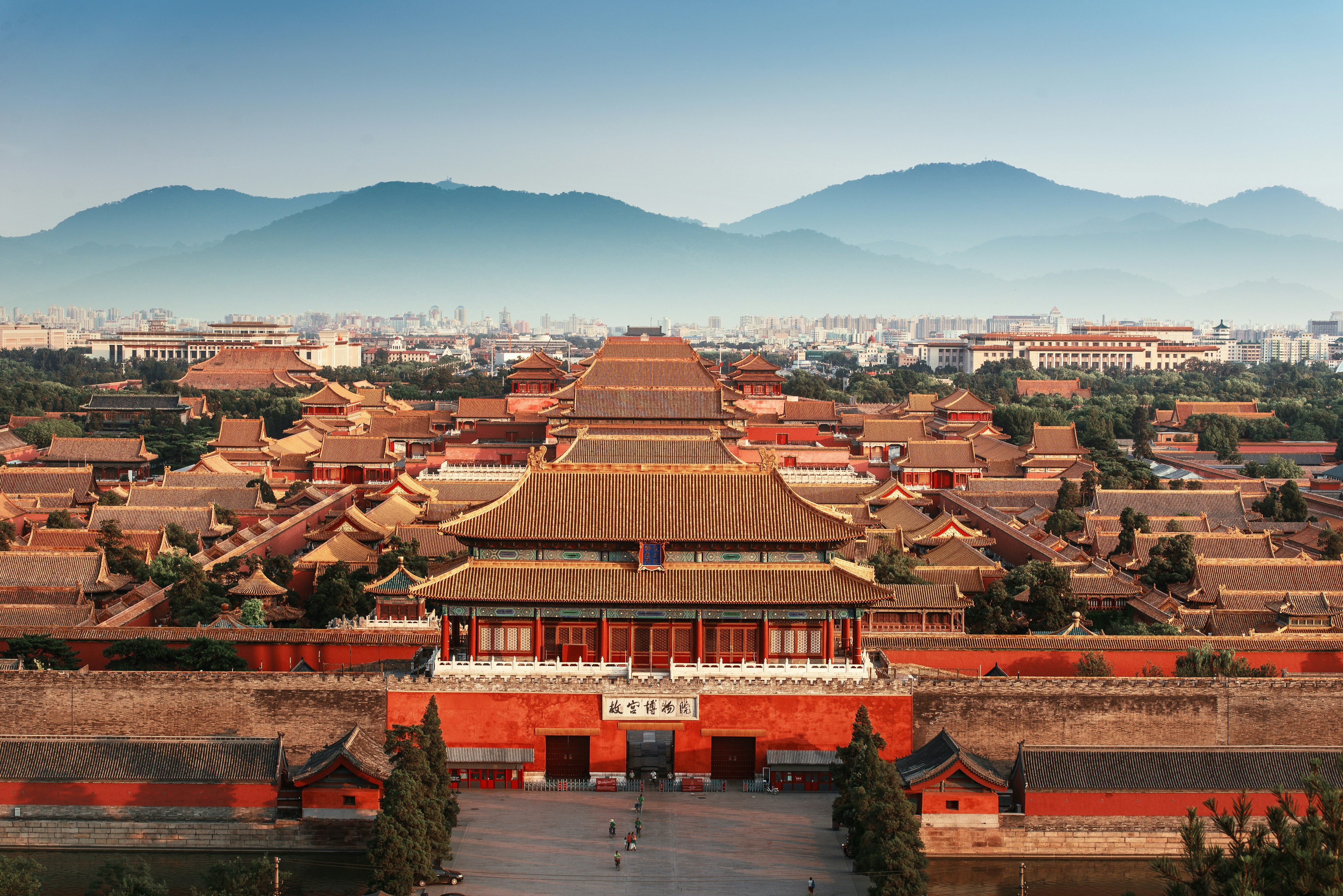 Aerial view of the Forbidden City in Beijing, China. The image shows the sprawling complex of traditional Chinese imperial buildings with golden roofs and red walls. In the foreground is the main entrance gate with a large courtyard. The city skyline and misty mountains are visible in the background, creating a striking contrast between ancient architecture and modern urban landscape.