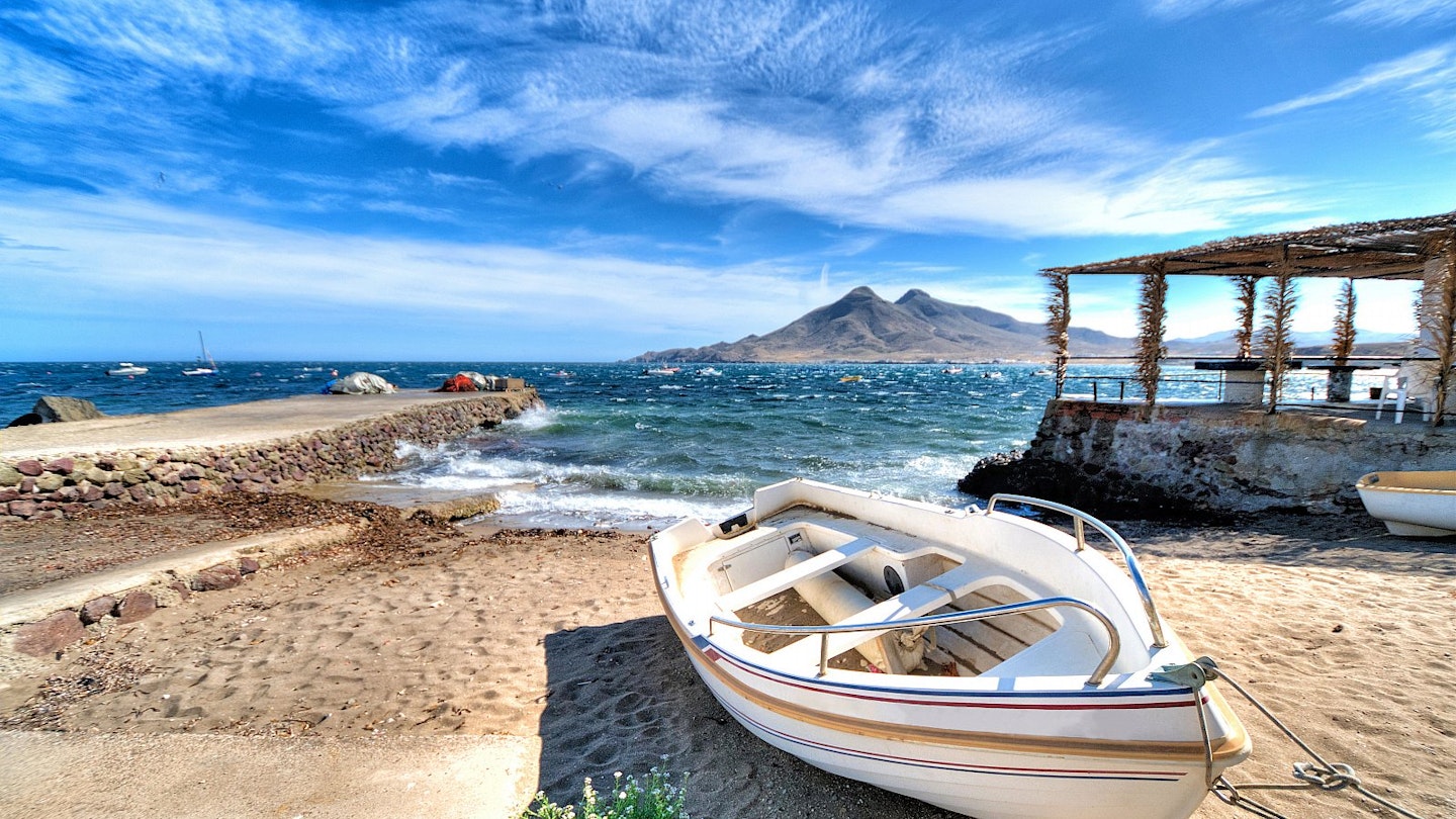 A white boat moored on a beach, with a jetty to the left and a stone structure to the right; out to sea is a dramatic peaked island.